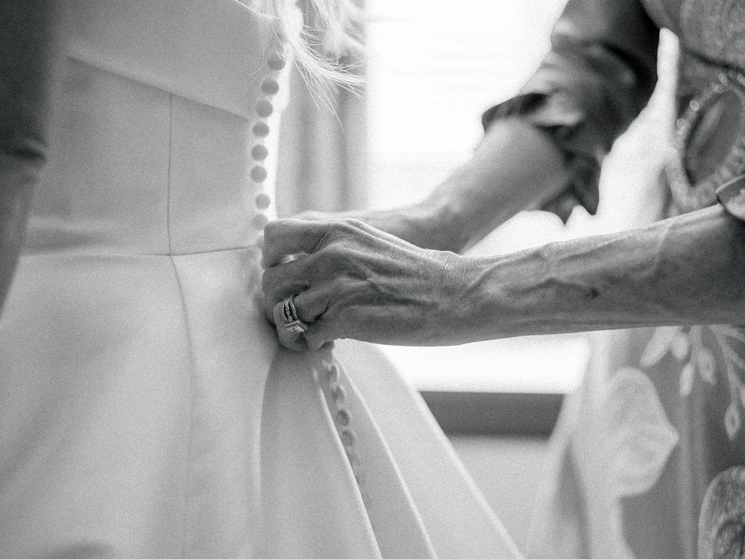 Close-up of a person's hand fastening buttons on clothing, wearing a ring on the finger, with other clothing and objects in the background.