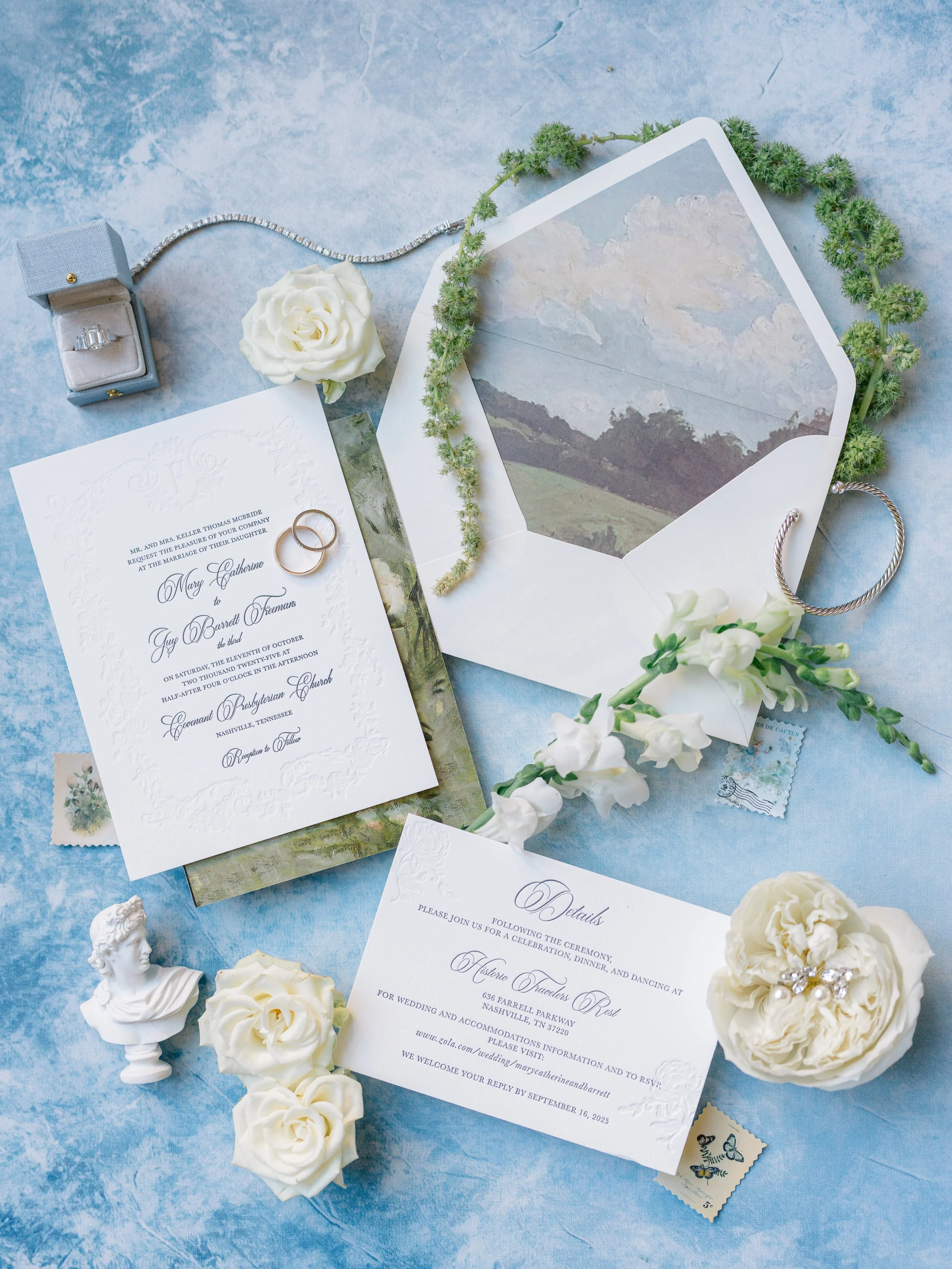 Wedding invitation with rings, flowers, and a necklace on a blue surface.
