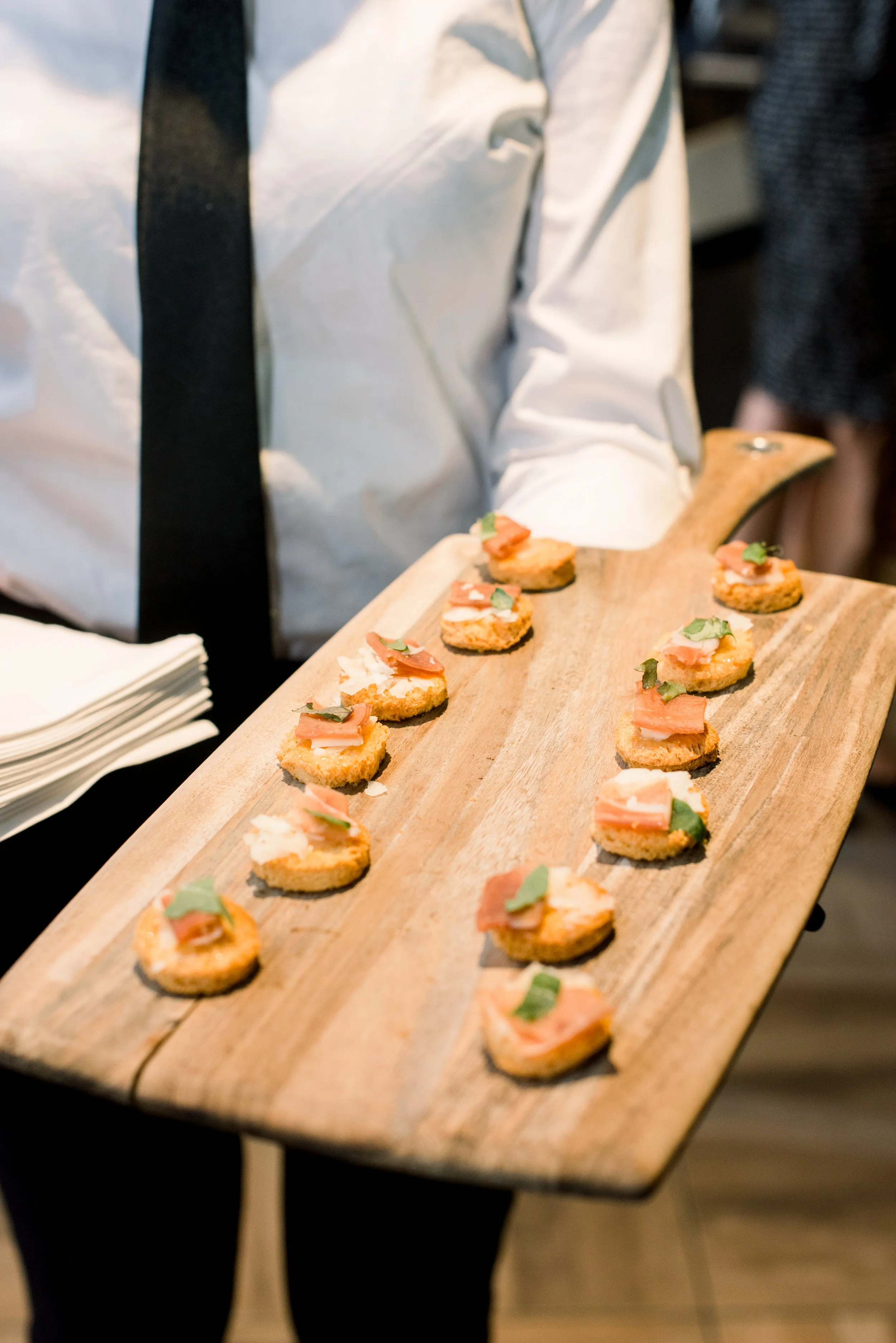 Person holding a wooden serving tray with small appetizers topped with cured meat and herbs at a gathering.