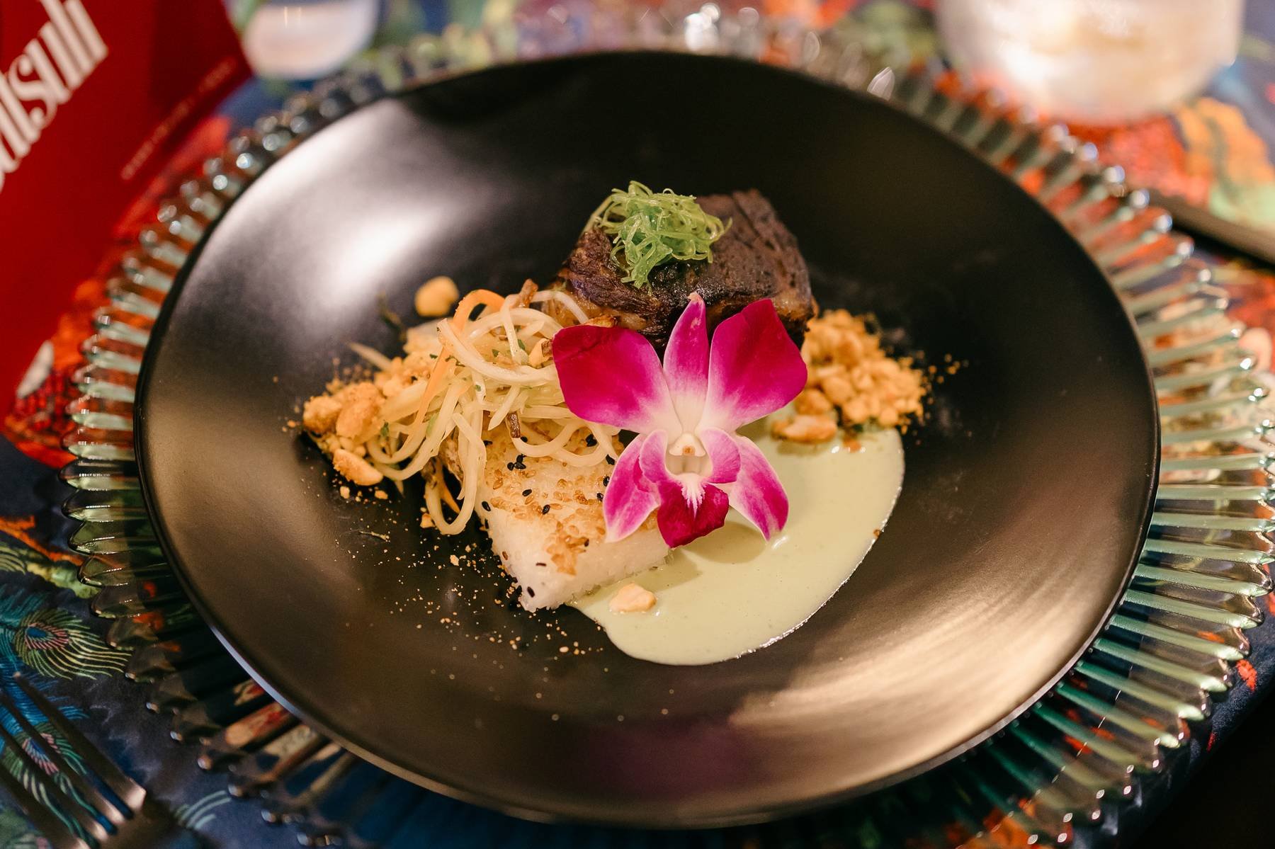 A black bowl with an elegant dish featuring a piece of fish topped with shredded scallions and a beef cube, garnished with a pink orchid flower, served with a green sauce and crunchy toppings, placed on a decorative glass plate.