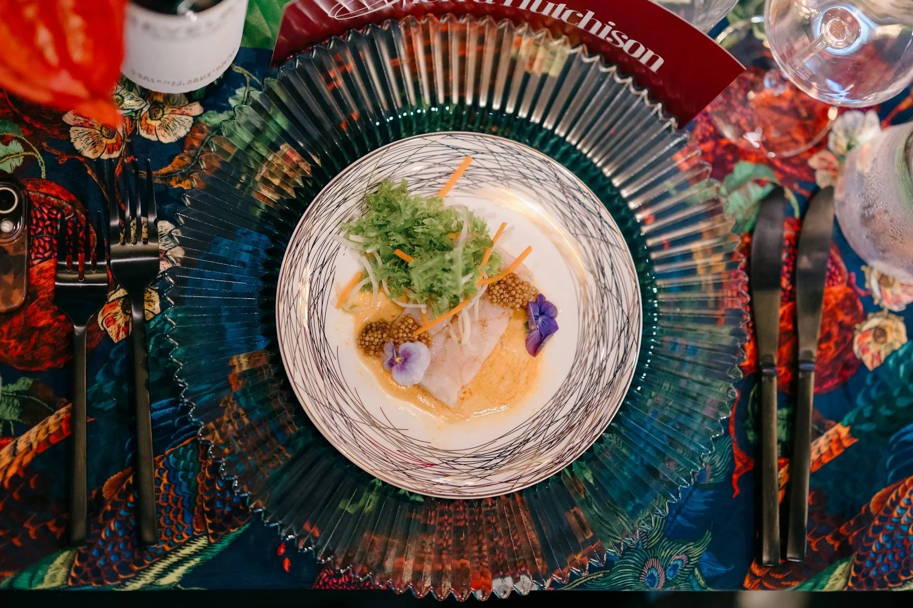 A plated dish with seafood garnished with herbs and edible flowers, set on a decorative tablecloth with cutlery and glasses surrounding it.