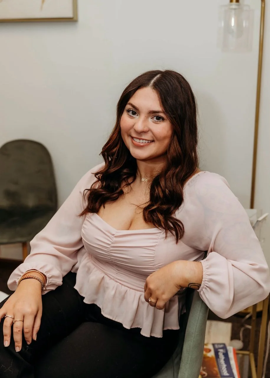 A woman with long, wavy brown hair smiling at the camera, sitting in a chair in an indoor setting.