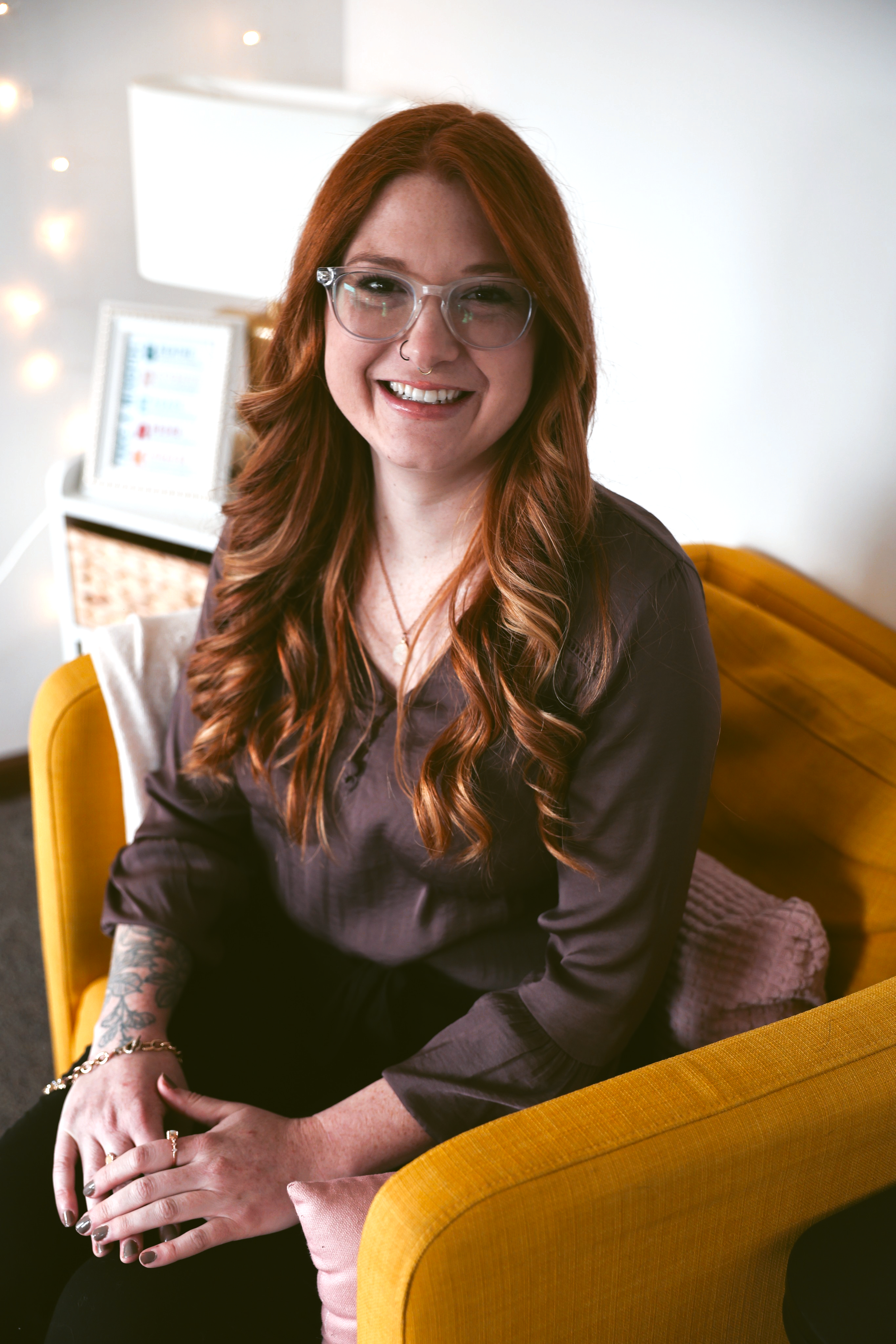 A woman with long red hair, glasses, and a nose ring seated on a yellow chair, smiling and looking at the camera, in a brightly lit room with a white wall and a framed picture in the background.