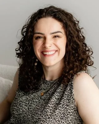 Smiling woman with curly dark hair, wearing a sleeveless animal print top and jewelry, sitting on a couch against a plain background.