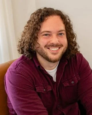 A man with curly hair and a beard smiling, wearing a burgundy shirt, sitting indoors.