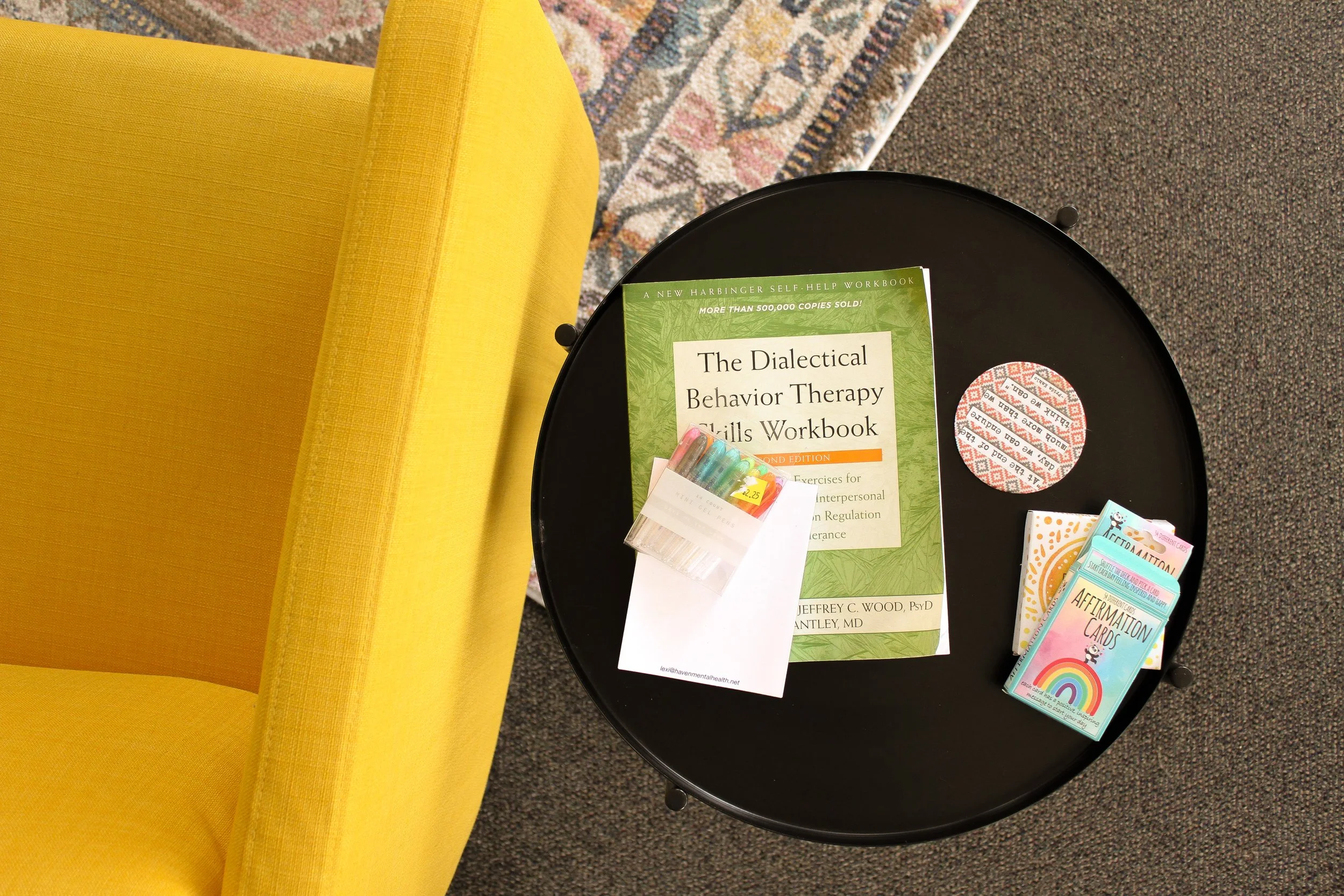 A black round side table holding a book titled 'The Dialectical Behavior Therapy Skills Workbook' and a small pack of colorful highlighters. There is also a box of affirmation cards and a store sticker on the book. A yellow upholstered chair is adjacent, and a colorful patterned rug is partially visible on the floor.