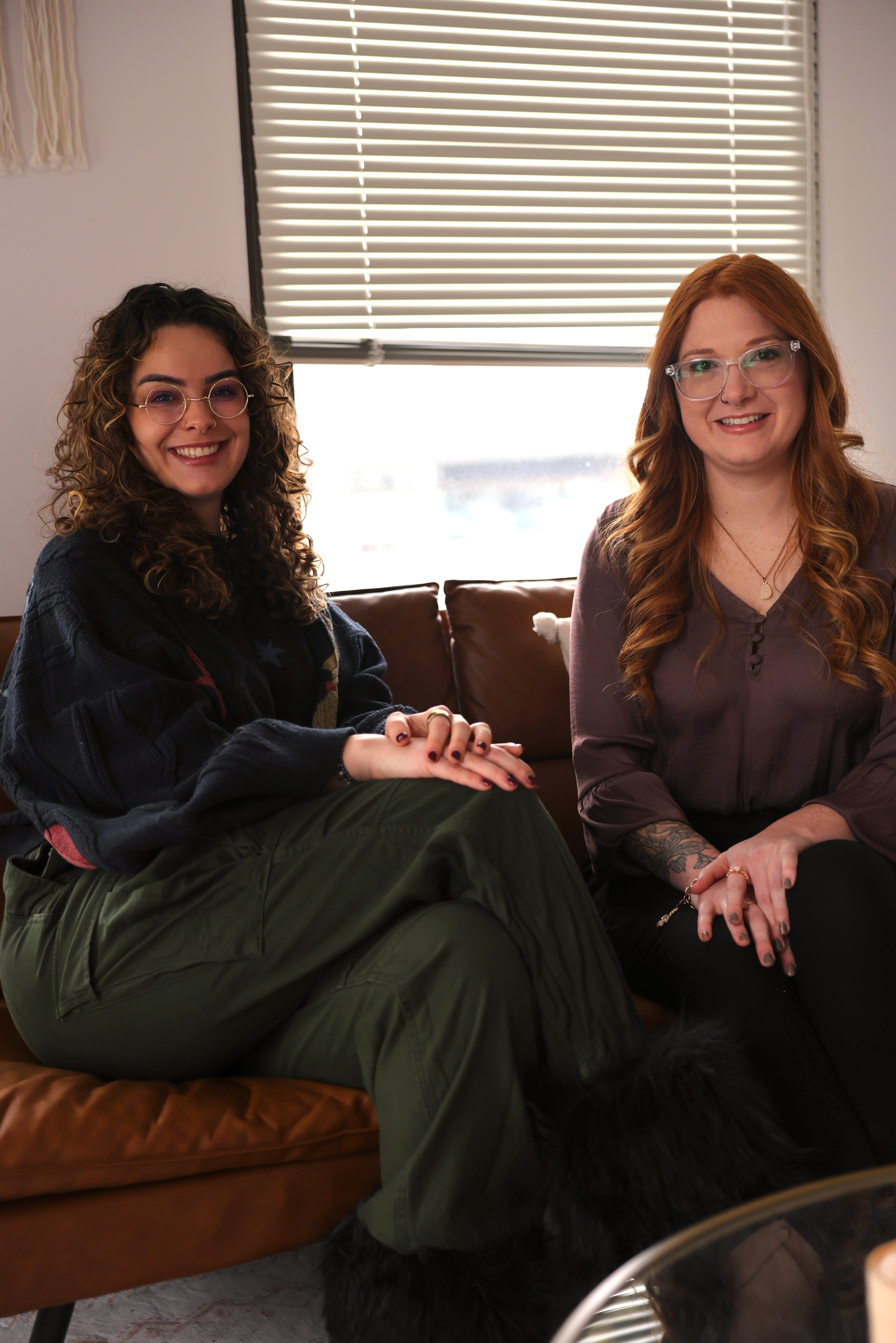 Two women sitting on a brown leather couch in a room with blinds on the window behind them, smiling at the camera.