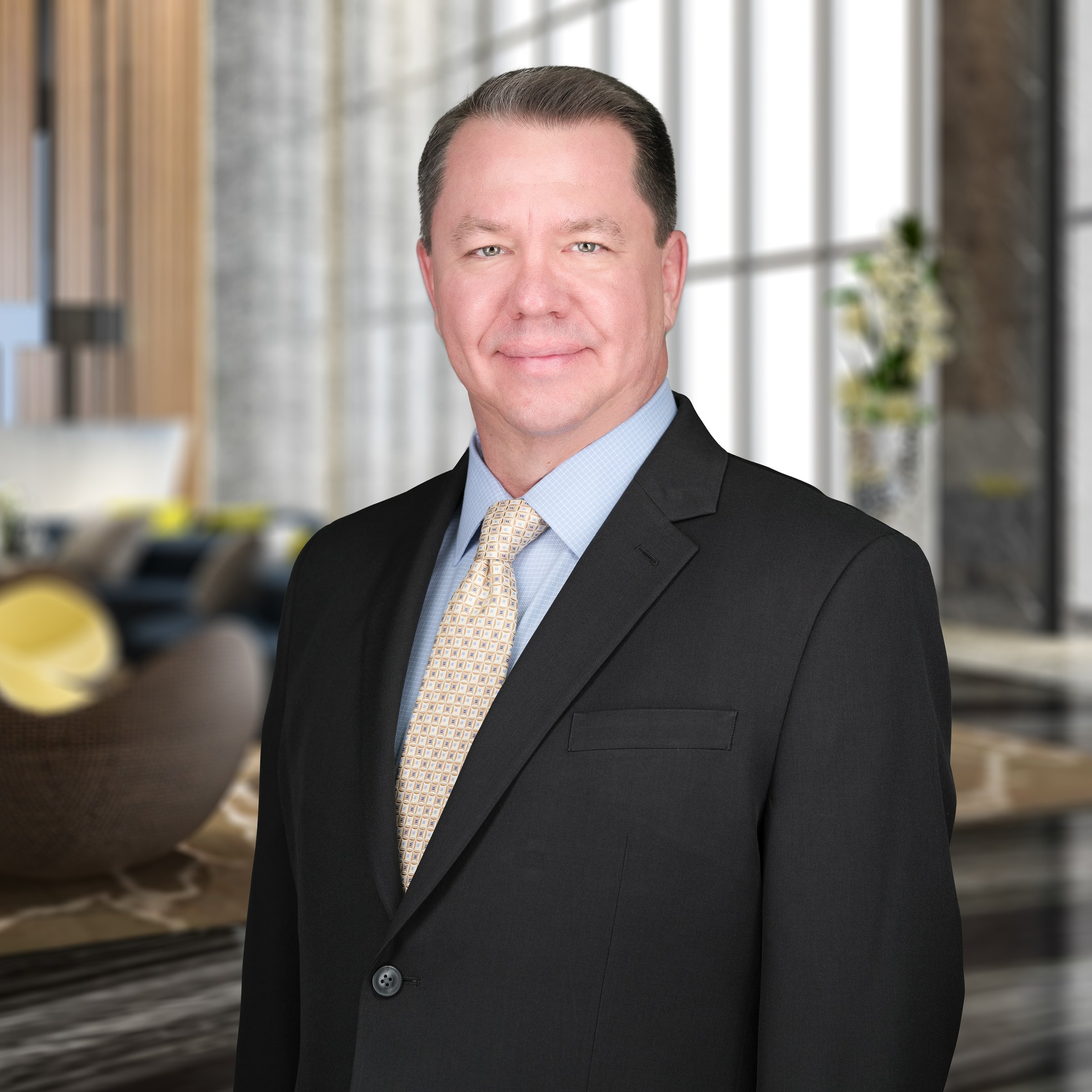Man with curly hair, glasses, and a beard, wearing a dark green sweater, sitting indoors with a plant and wall in the background.