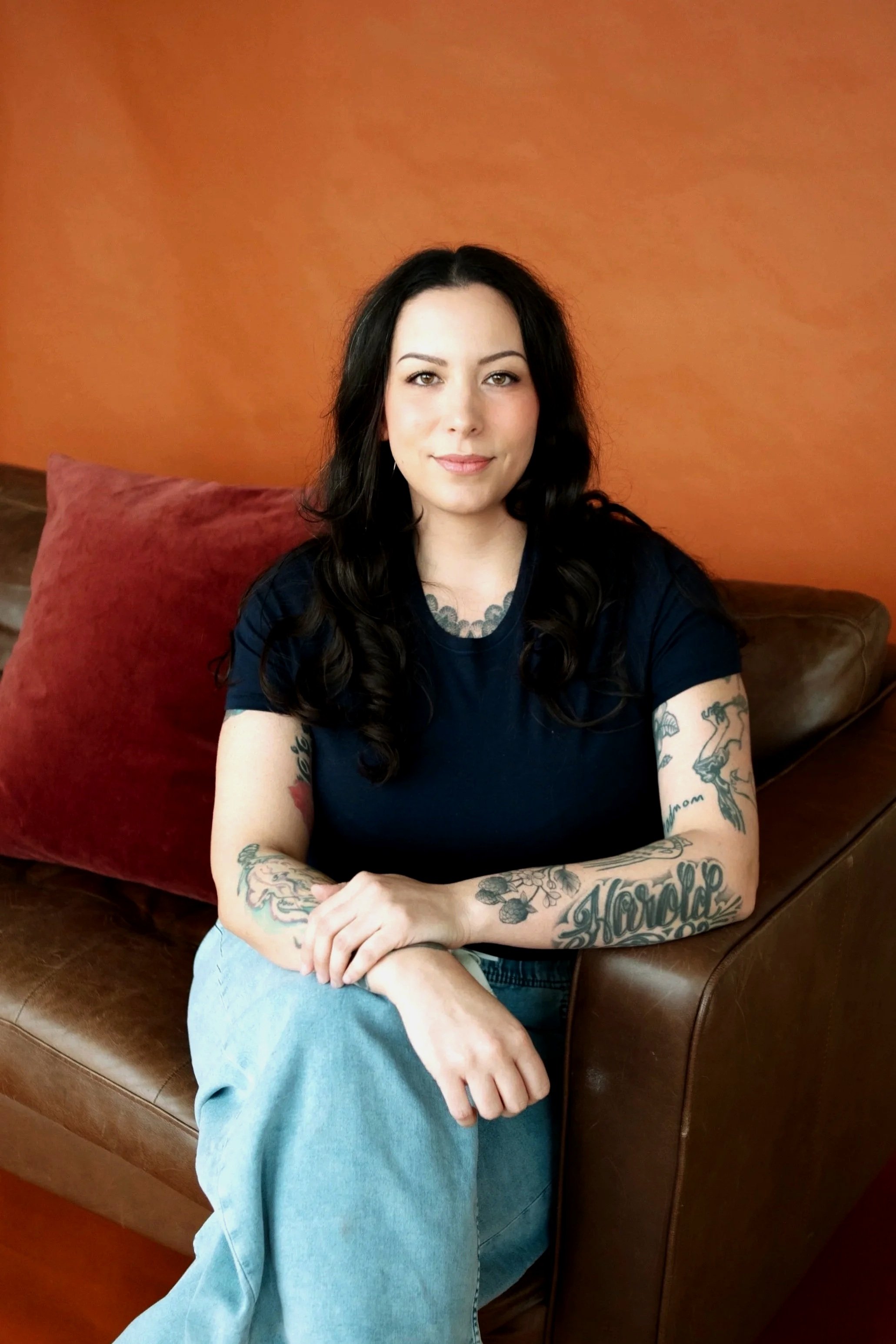 A woman with black hair, tattoos on her arms, and wearing a black shirt and blue jeans, sitting on a brown leather couch with an orange wall in the background.