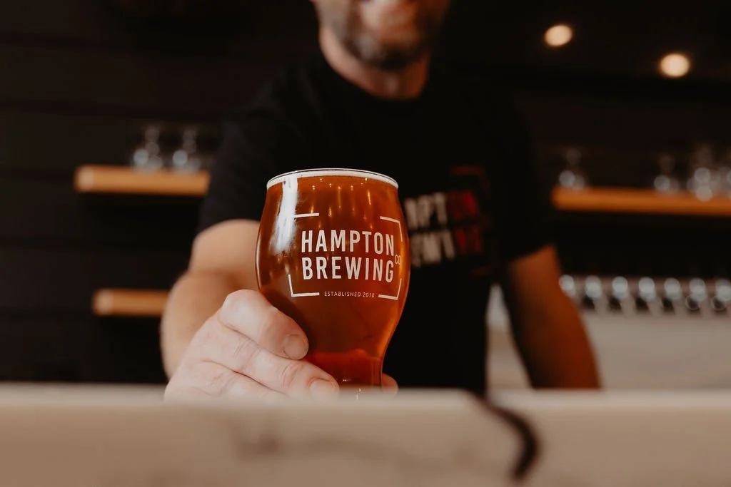 A person holding a glass of amber-colored beer with a Hampton Brewing logo, in a bar or brewery setting.