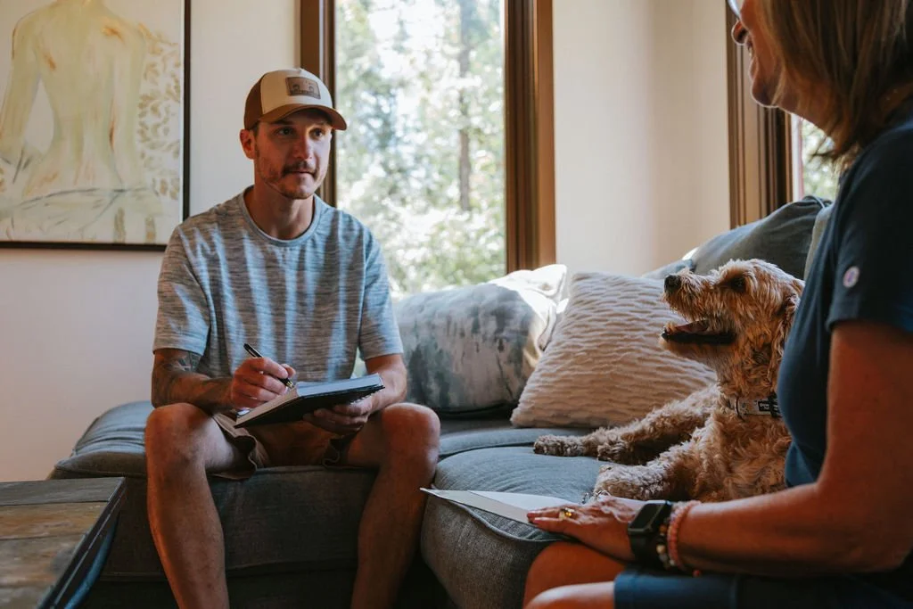 A man in a gray t-shirt and a cap listens to a woman with a dog during a counseling or therapy session in a living room with large windows and natural light.