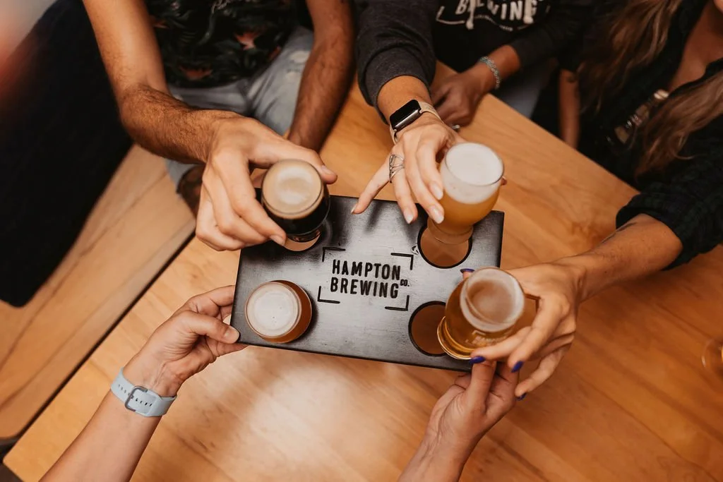 Four people clinking beer glasses over a wooden table with a black flight tray labeled "Hampton Brewing Co."