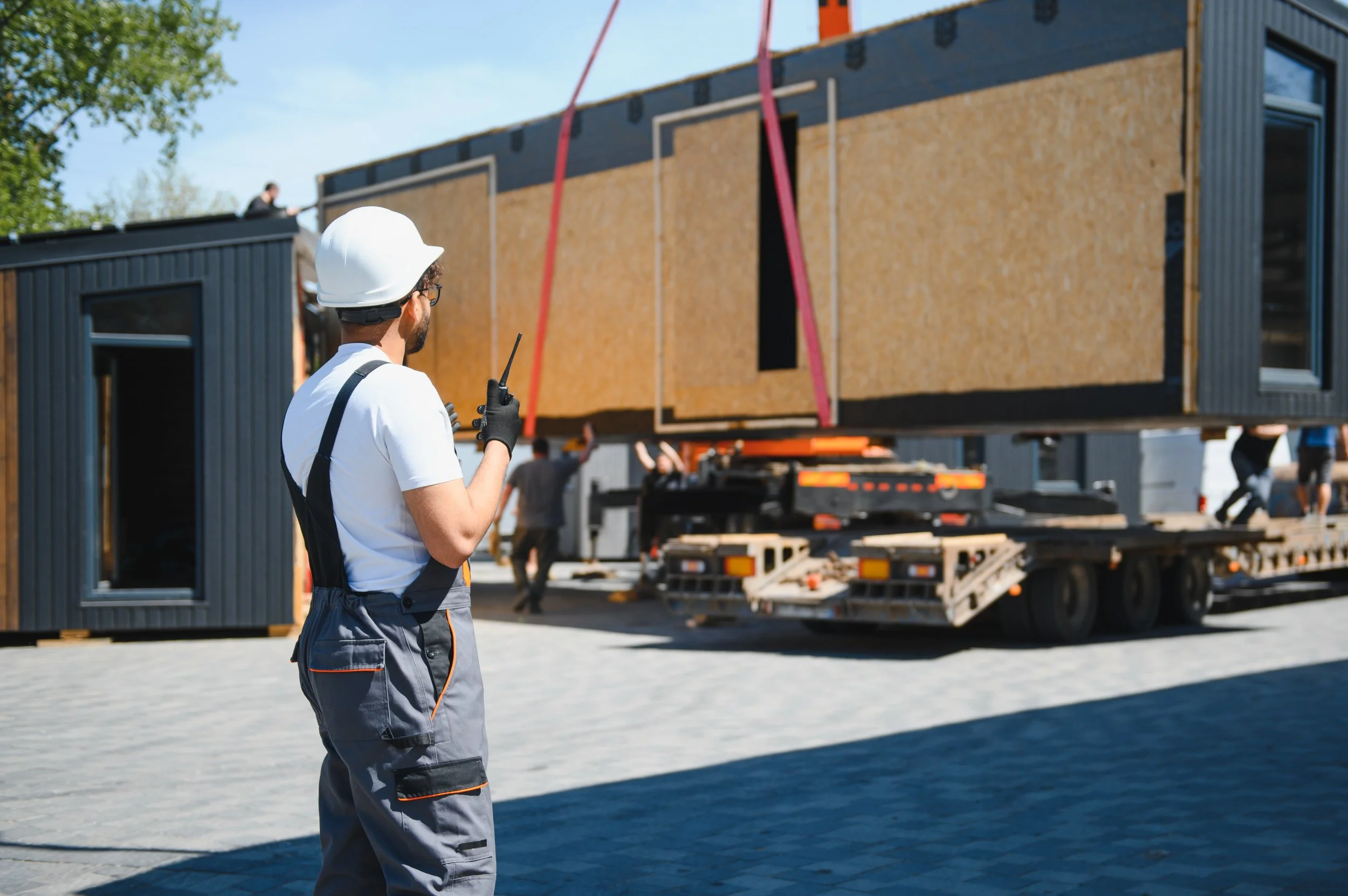 A construction worker wearing a white safety helmet, glasses, a white t-shirt, and gray work pants, standing outside near a body of water, holding a walkie-talkie, with a house under construction on a trailer in the background.