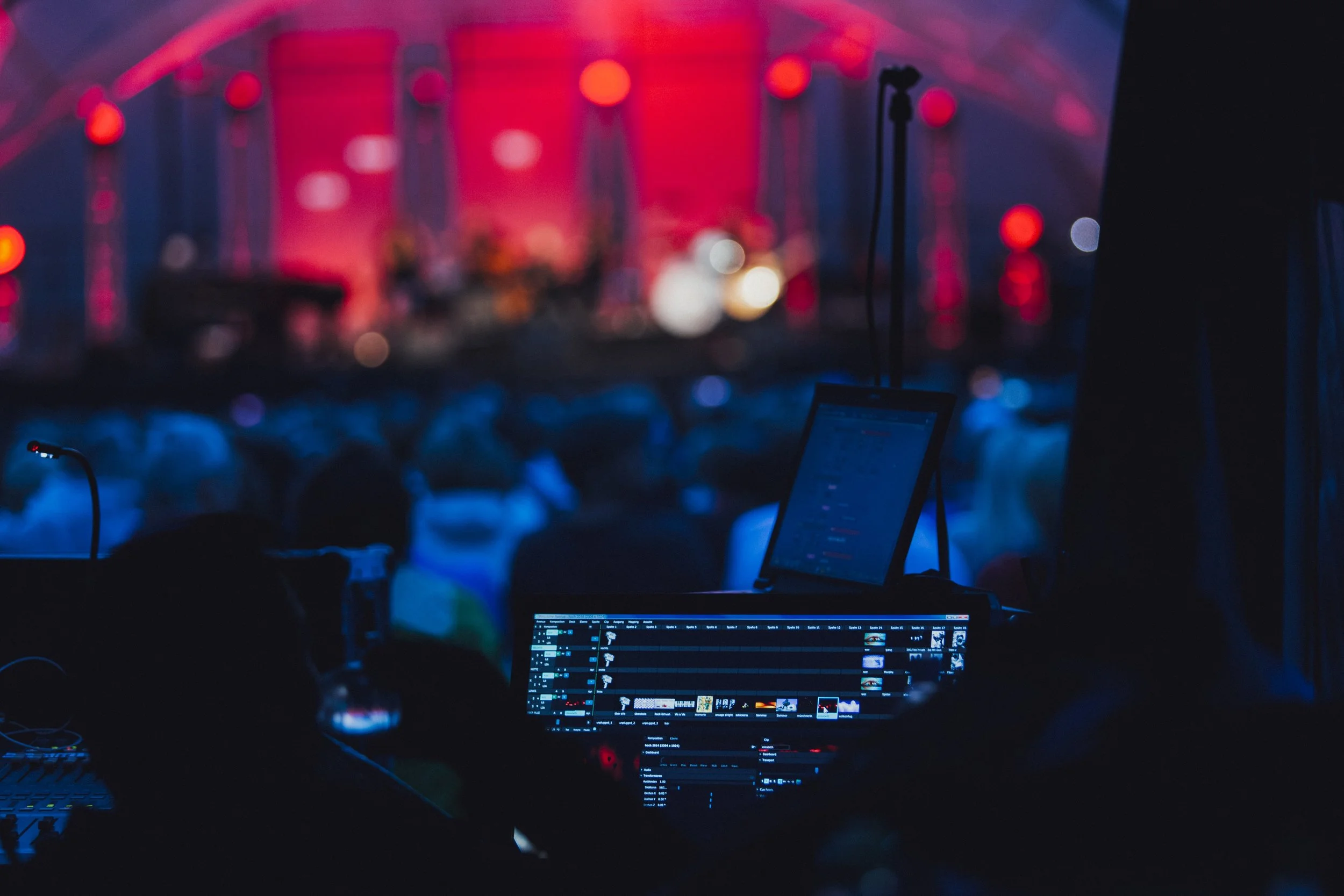 A dark room with DJ equipment and a laptop displaying audio editing software, illuminated by colorful stage lights with a pink and blue glow.