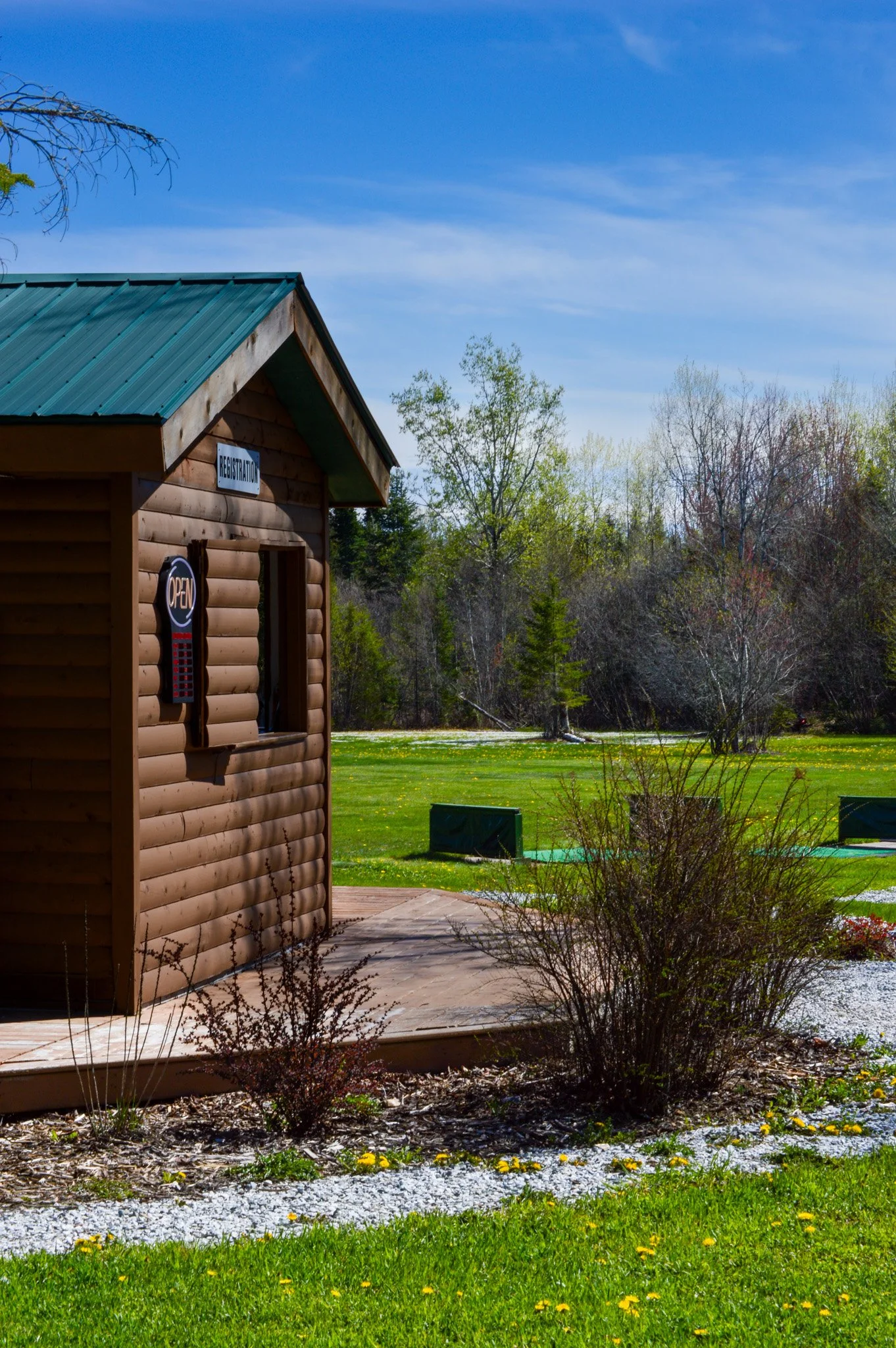 A small wooden building with a teal metal roof, a sign that says 'Registration,' and an 'Open' sign on the window. The building is surrounded by green grass, bushes, and trees under a blue sky with some clouds.