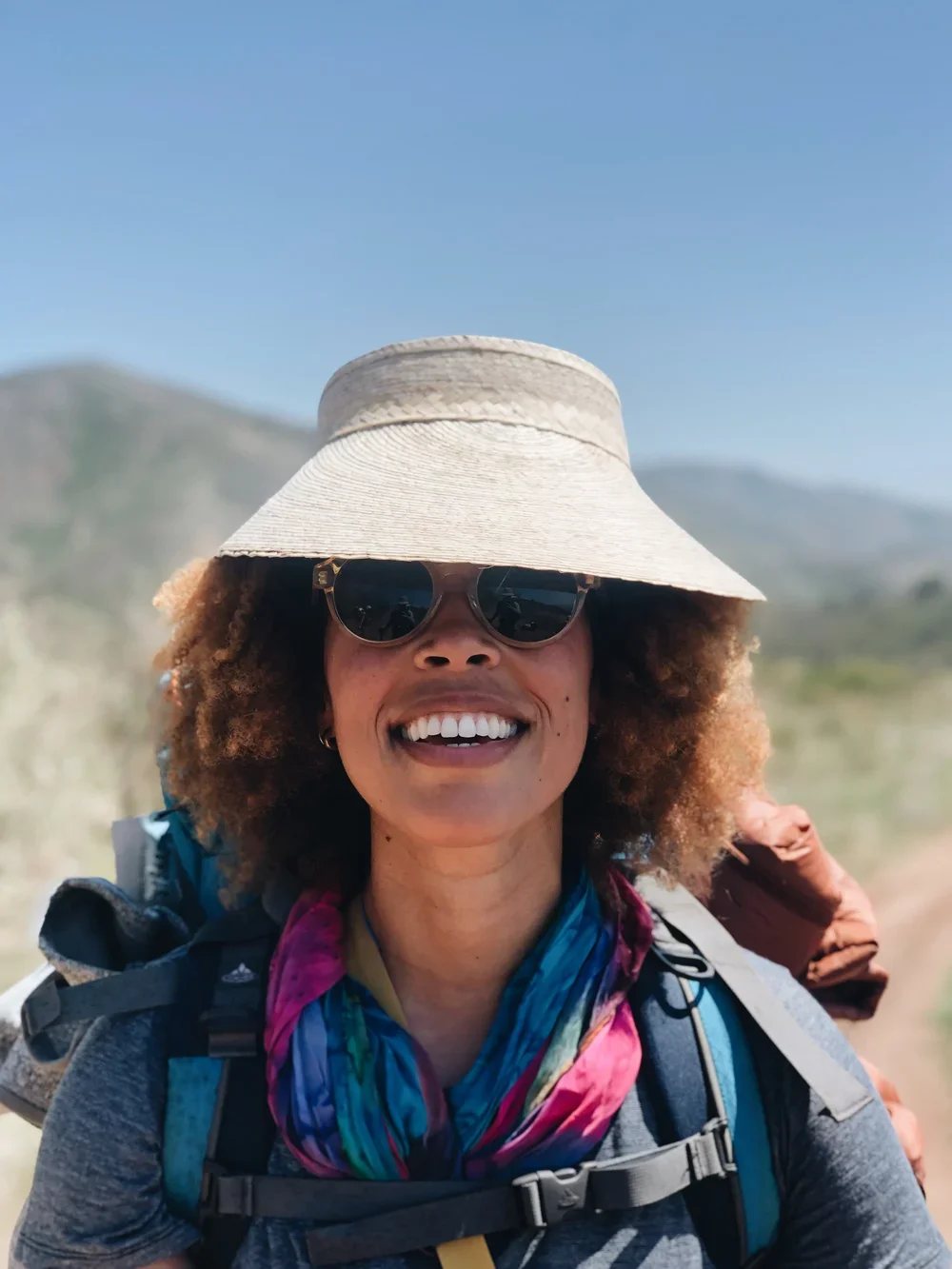 A woman with curly hair wearing a wide-brimmed hat, sunglasses, a colorful scarf, and a backpack outdoors with mountains and blue sky in the background.