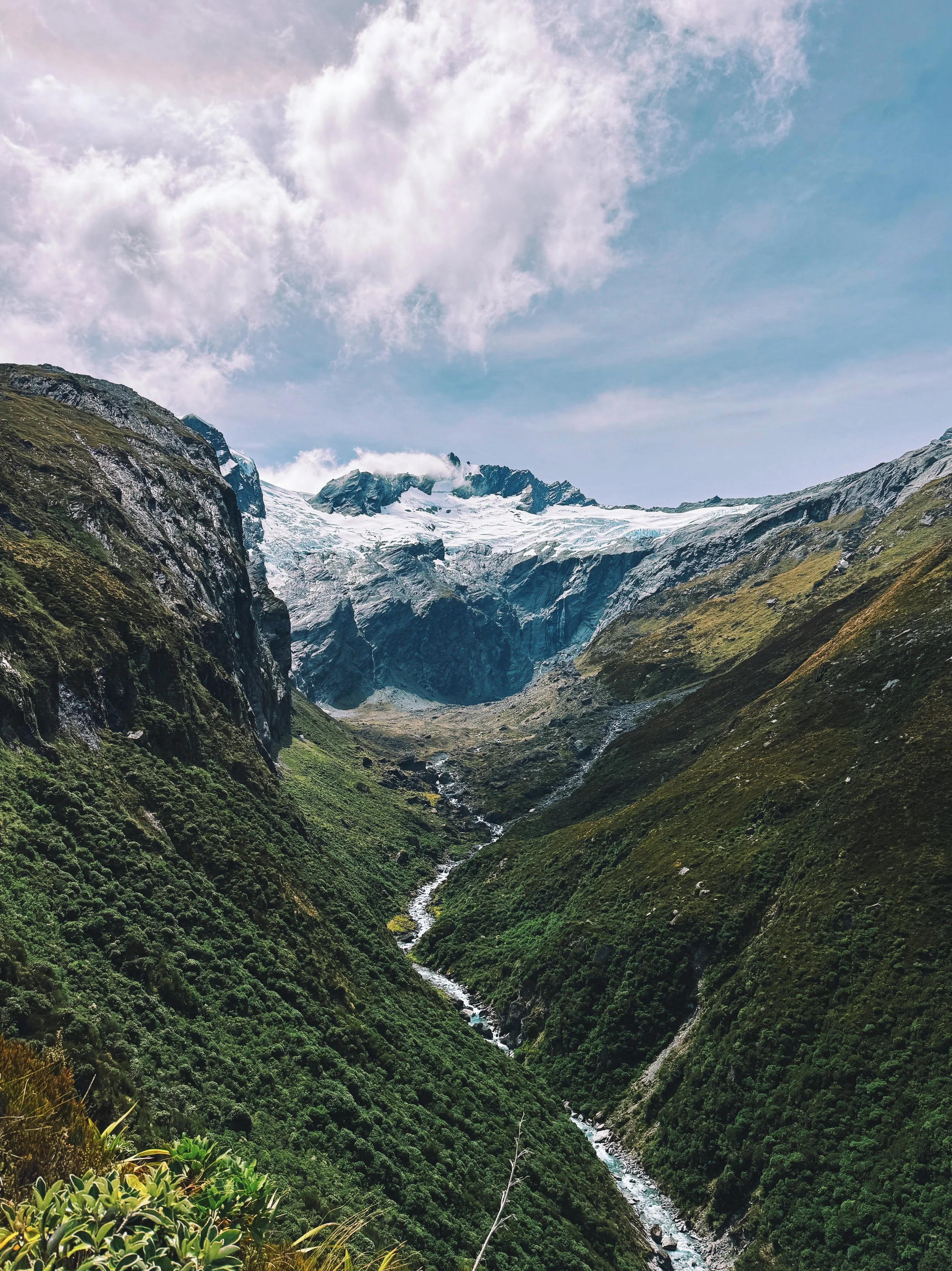 A mountain landscape with a valley, a small river flowing through green vegetation, and snow-covered peaks under a partly cloudy sky.