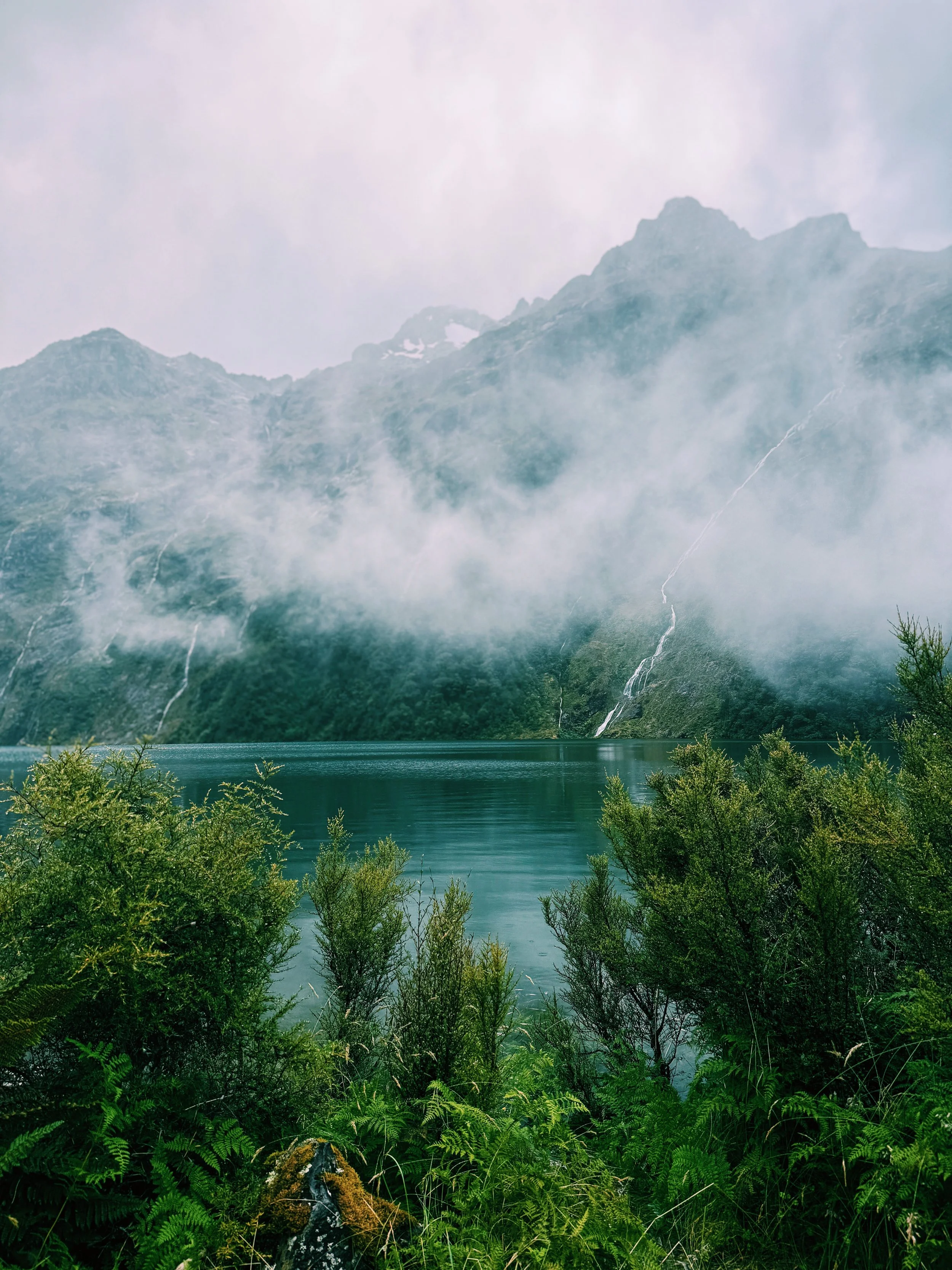 A mountain with multiple waterfalls cascading down into a lake, surrounded by lush green foliage in the foreground, with clouds or mist partially covering the mountain peaks.