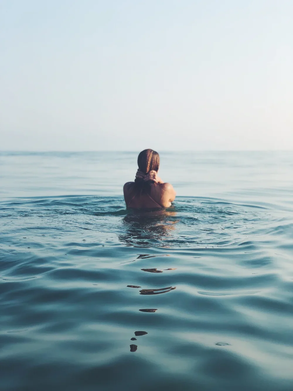 Person with long hair in the ocean, viewed from behind, looking towards the horizon.