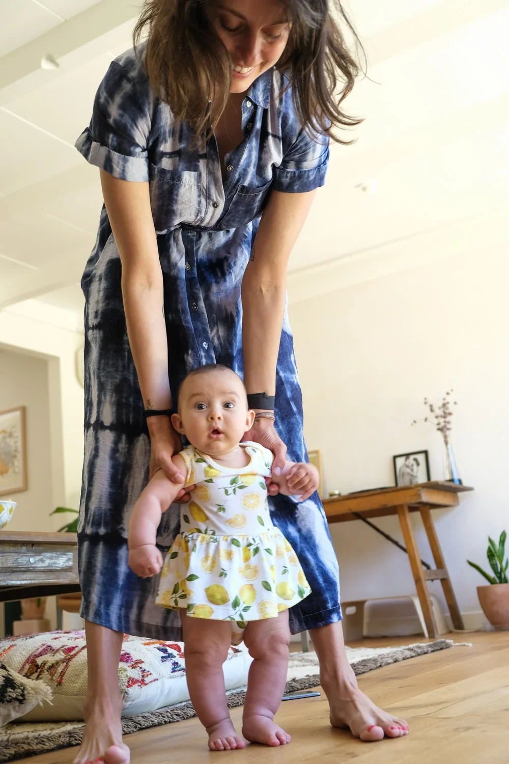 A woman in a blue tie-dye dress smiling and holding a toddler in a lemon-patterned dress inside a cozy home.