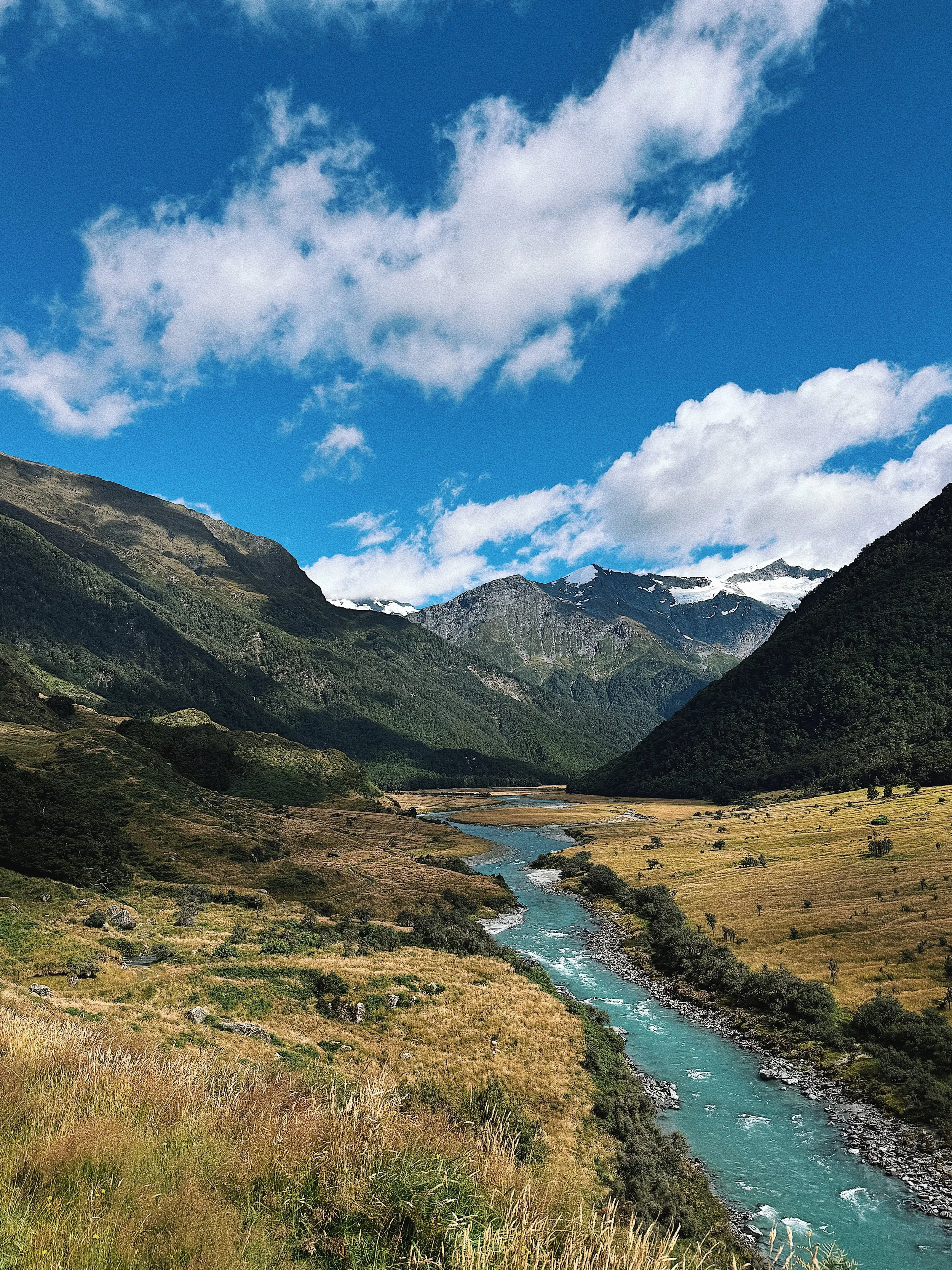 A scenic view of a mountain valley with a winding river, green hills, snow-capped mountains in the background, and a partly cloudy blue sky.