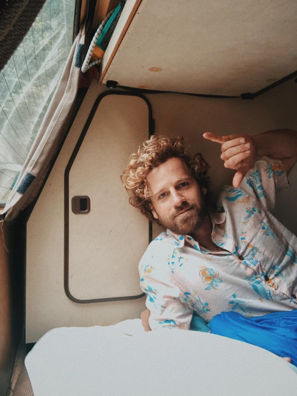 A man with curly blond hair and a beard making a hand gesture. He is lying on a bed in a room with an open window, wearing a Hawaiian shirt with floral patterns.