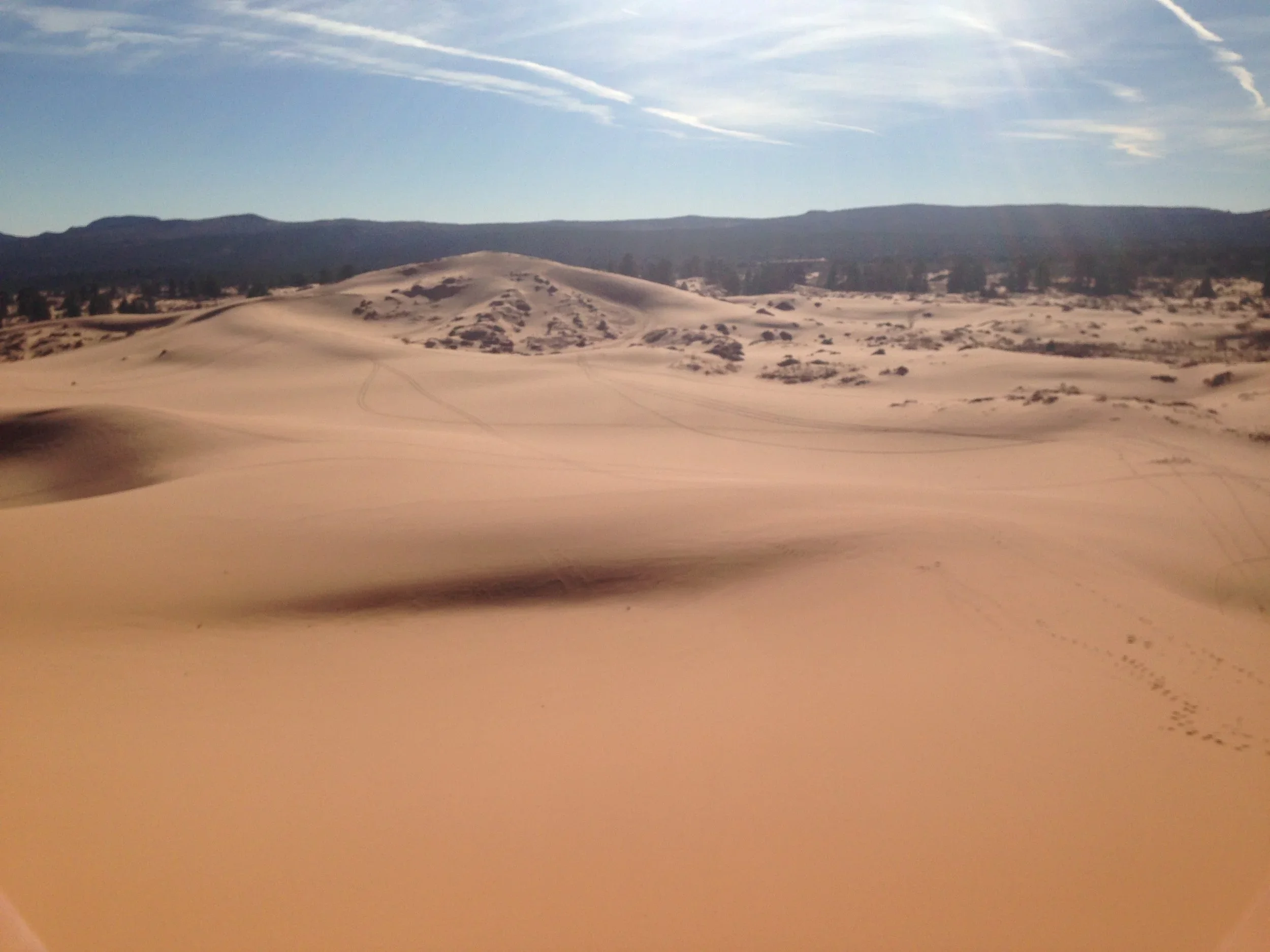 Photo of sand dunes with some tracks, distant hills, and a partly cloudy sky.