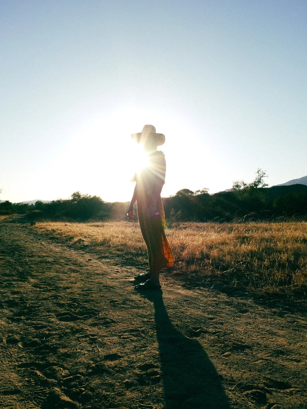 Person standing on a dirt path in a rural area with mountains in the background, wearing a hat and colorful clothing, with the sun setting behind them, casting long shadows.