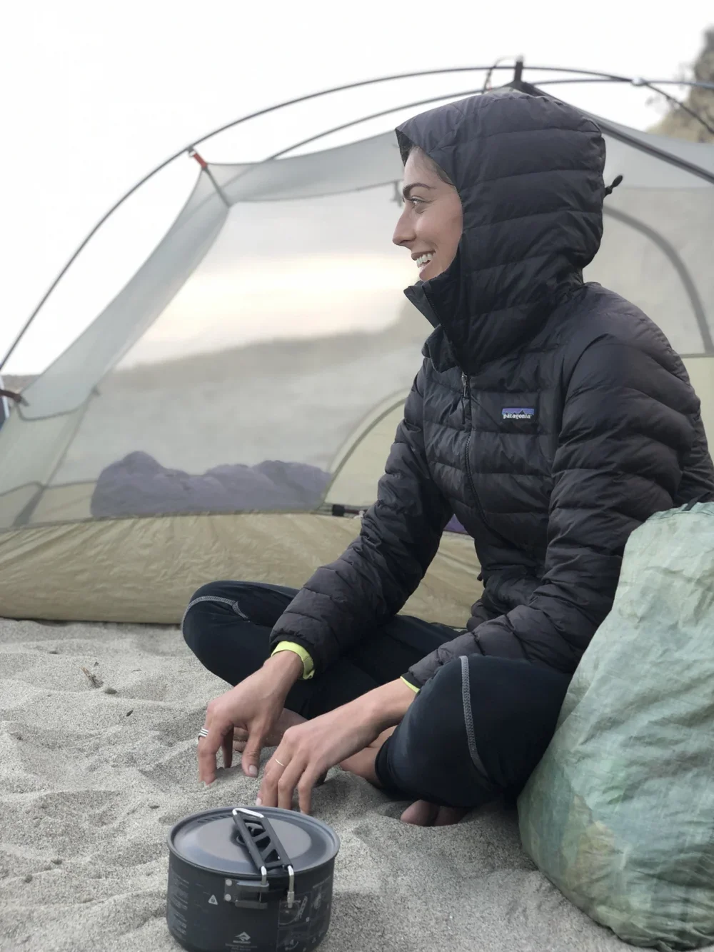 Woman in black jacket sitting on sand near a tent, smiling, with camping gear and a stove in front of her.