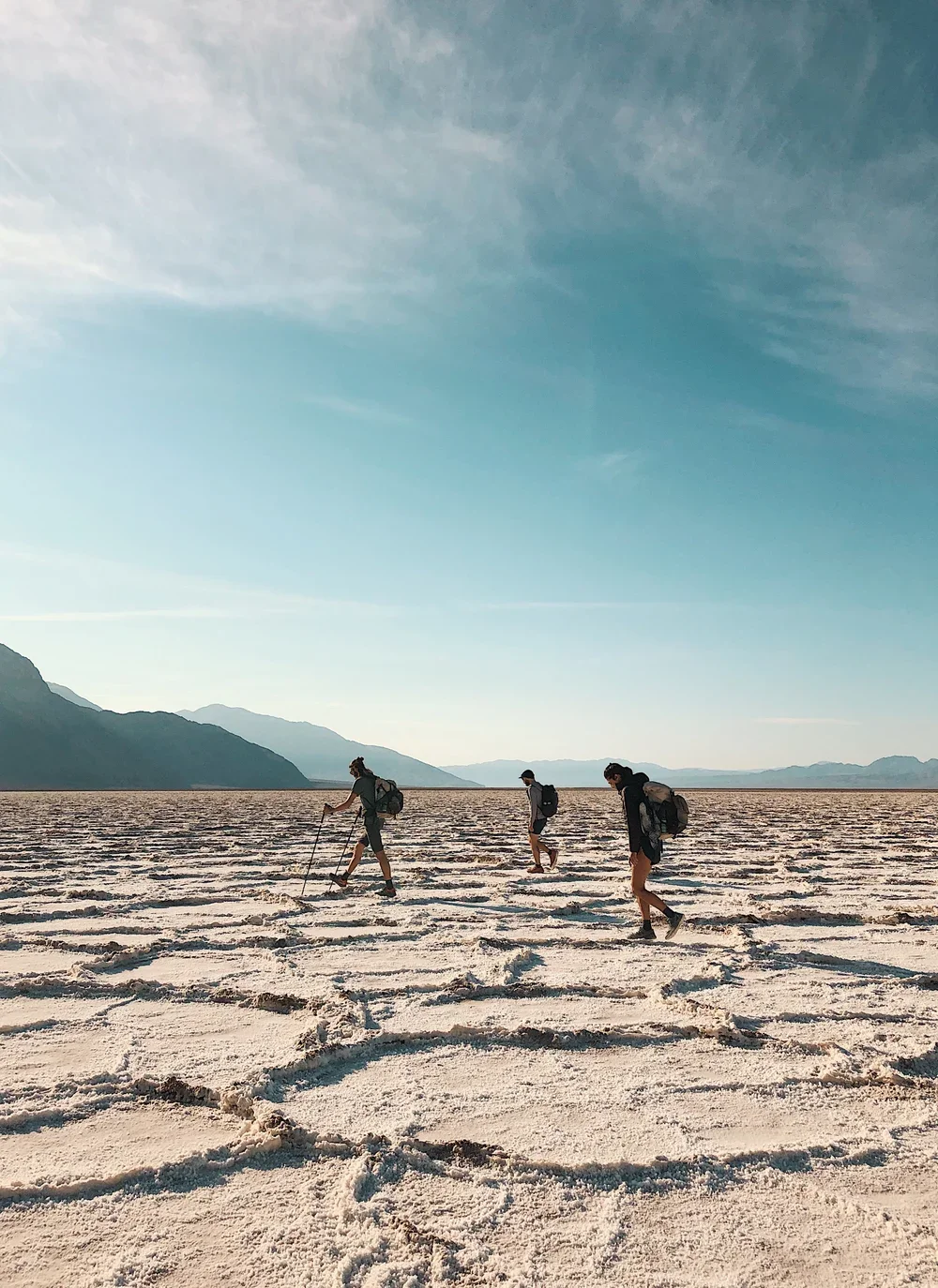 Three hikers with backpacks walking across a desert landscape with mountains in the background and a blue sky overhead.
