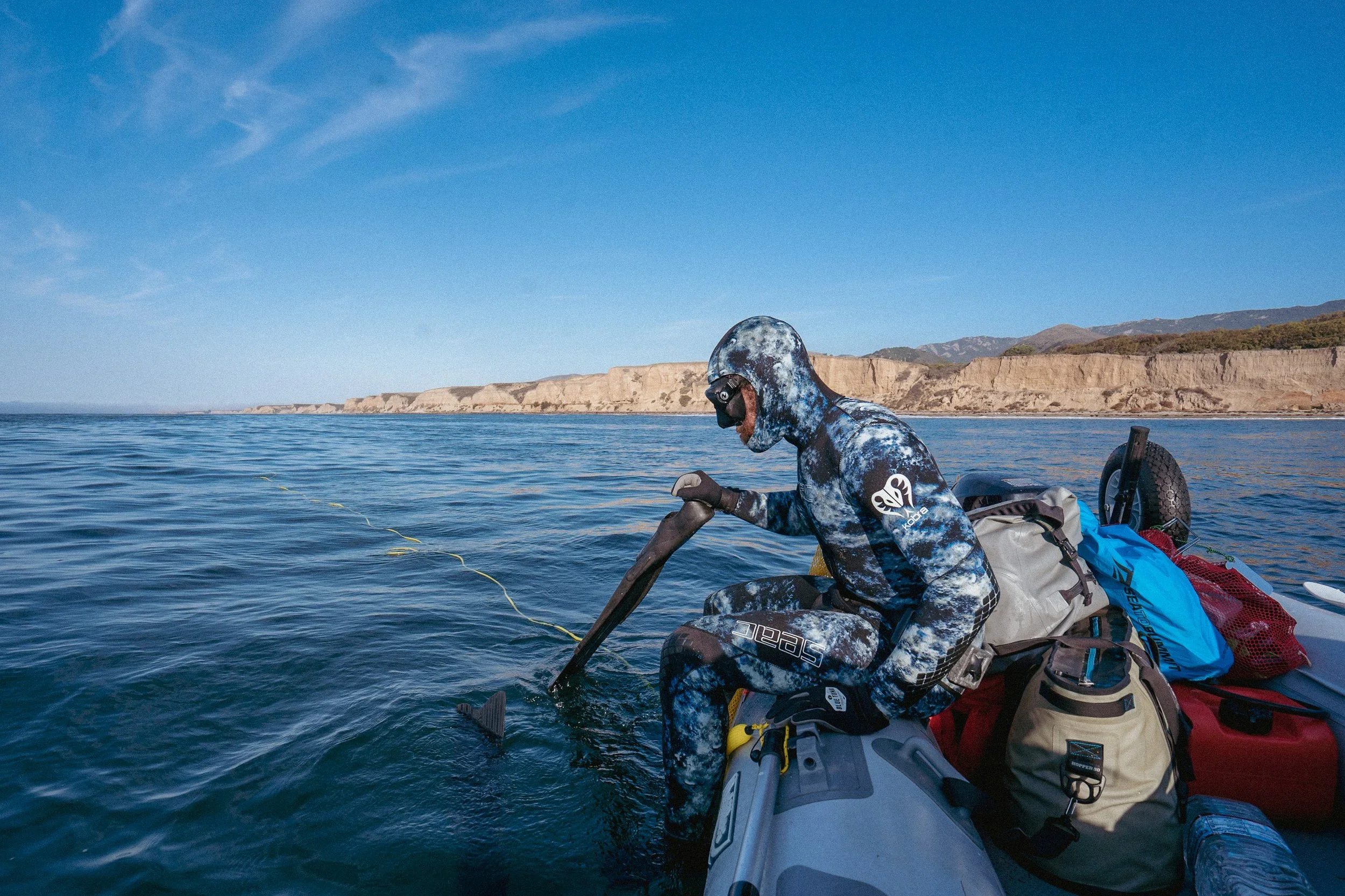 Person in a camouflage wetsuit and diving mask sitting in an inflatable boat, using a paddle to explore a calm body of water with a rocky shoreline in the background.