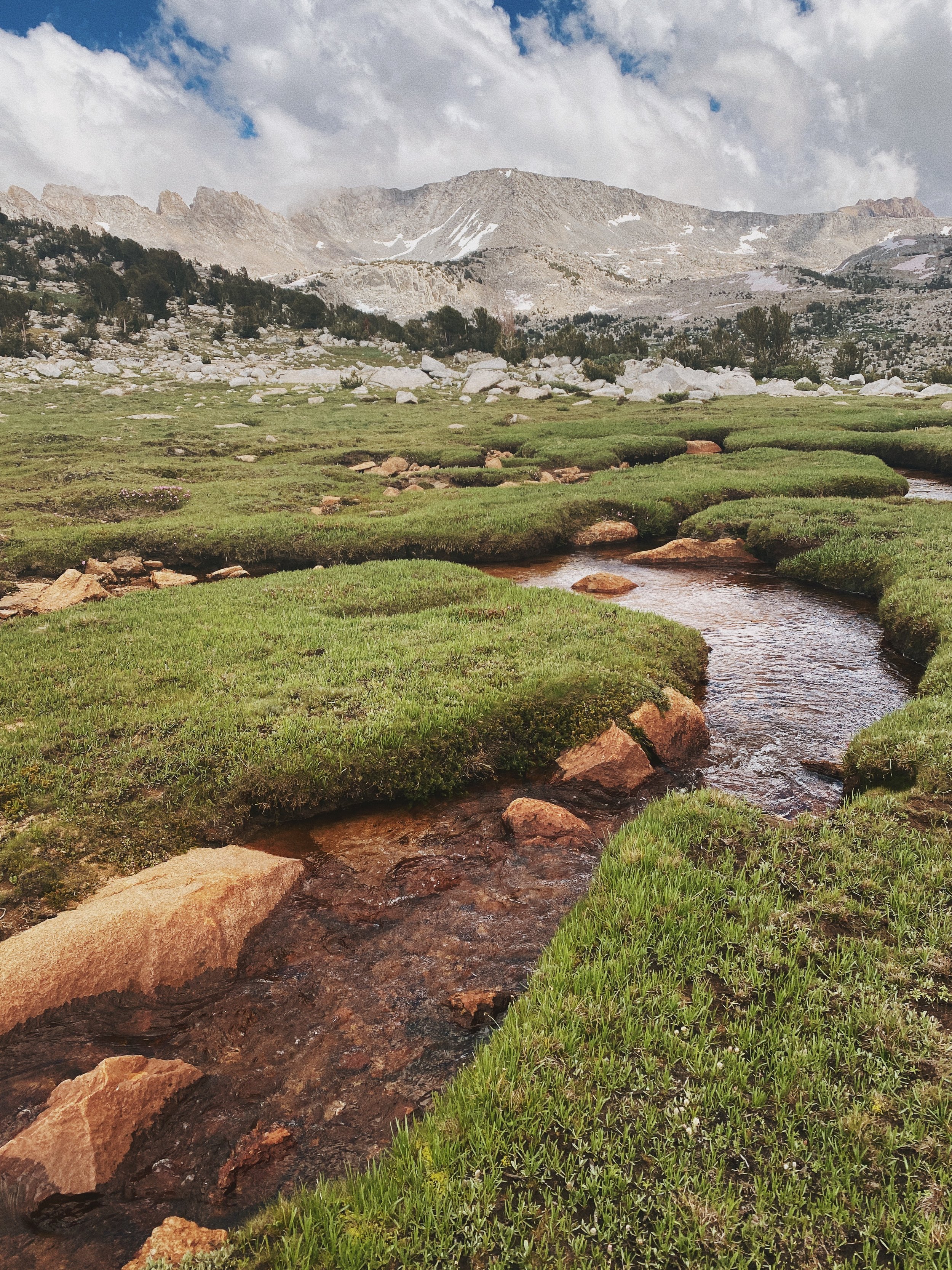 A mountain landscape with a meandering stream running through lush green grass, rocky terrain, and snow-capped peaks in the background under a partly cloudy sky.