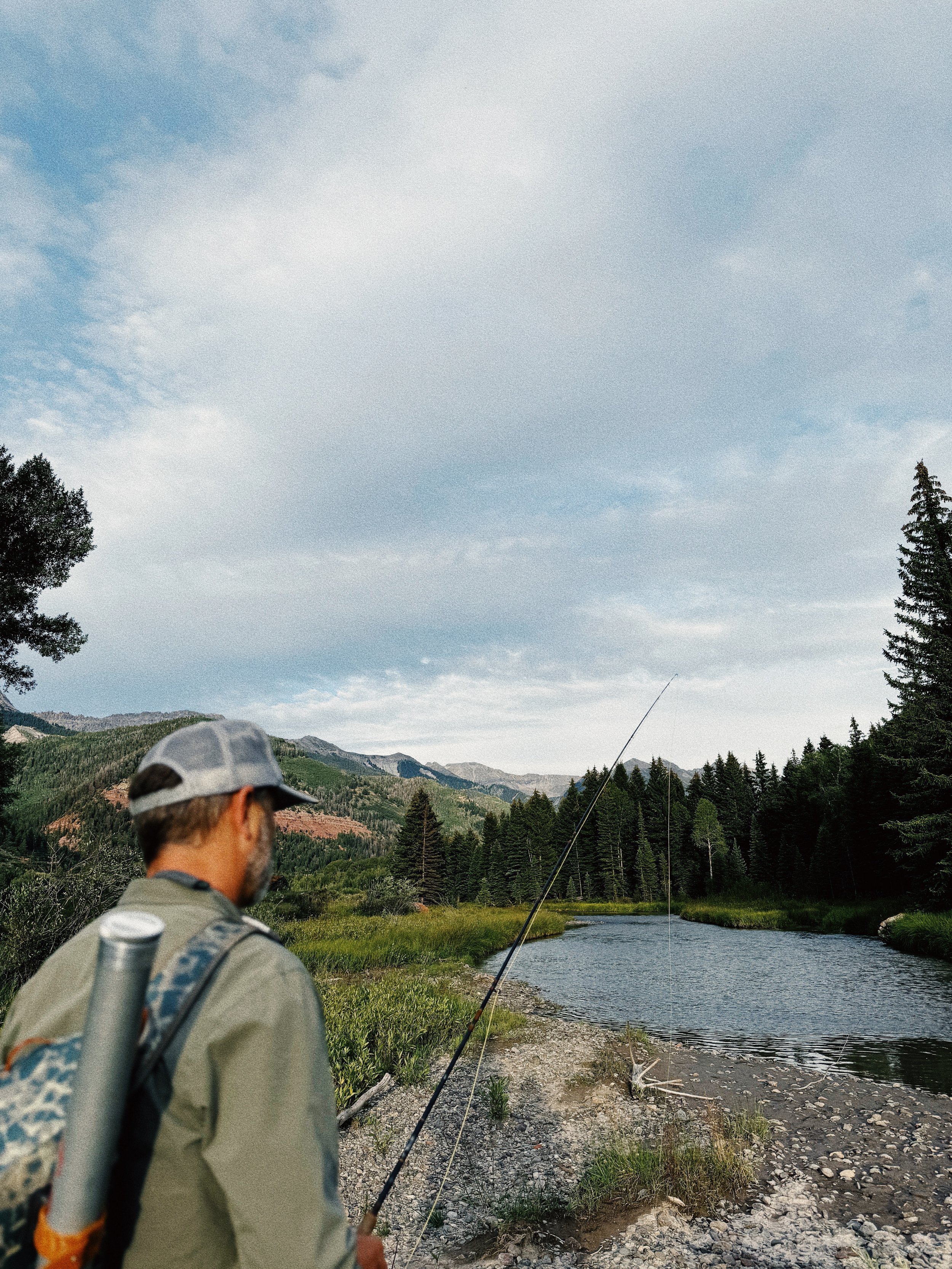 A man in a gray cap and outdoor clothing walking along a riverbank, carrying a fishing rod and a backpack, with a scenic landscape of trees, mountains, and a partly cloudy sky in the background.