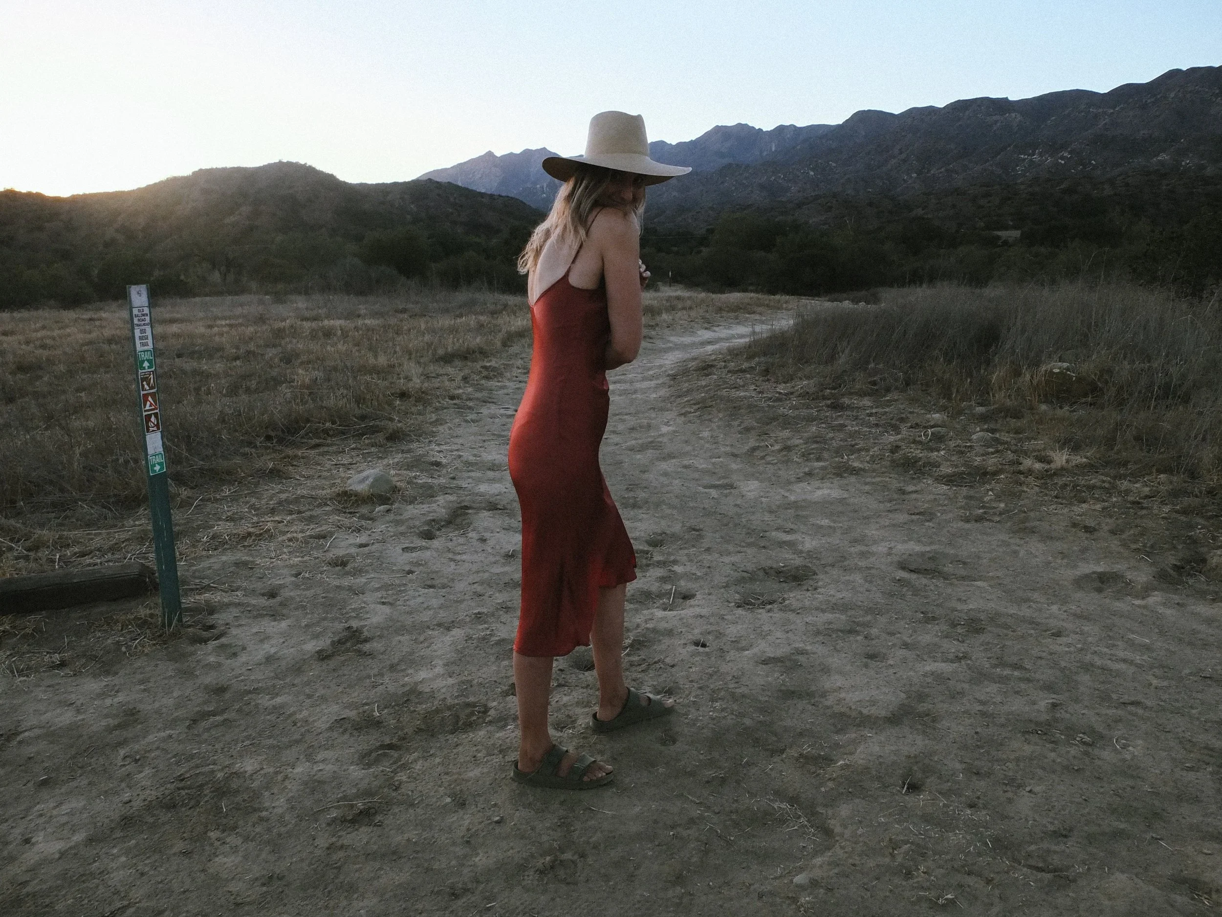 A woman in a red dress and wide-brimmed hat stands on a dirt trail in a field with mountains in the background, during sunset or dusk.