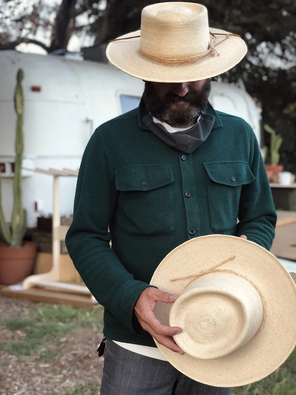 A man is holding a straw hat outdoors, wearing a large brimmed straw hat and a dark green jacket, with a blurred background of trees and a white trailer.