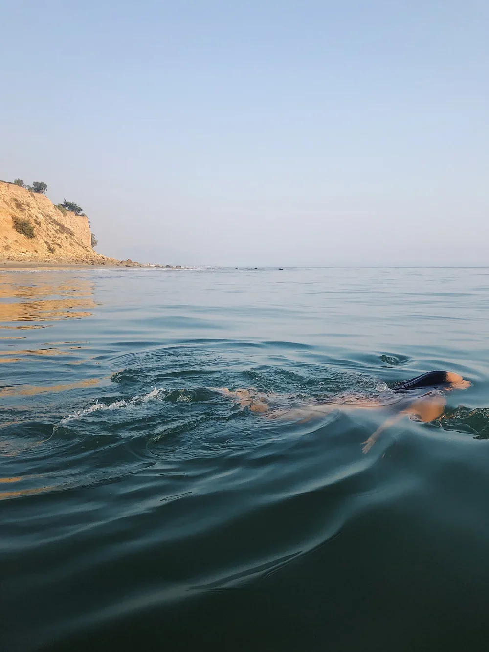 A person swimming in the ocean near a rocky coastline with cliffs.
