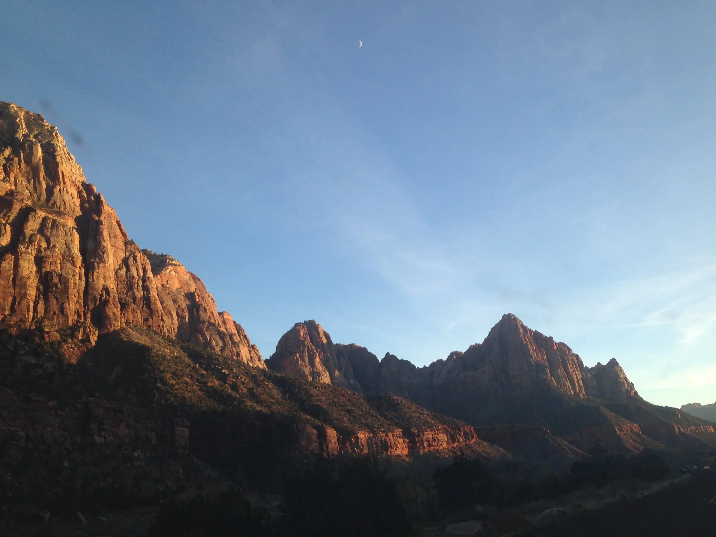 Sunset over rugged mountains with a clear blue sky and a visible crescent moon.