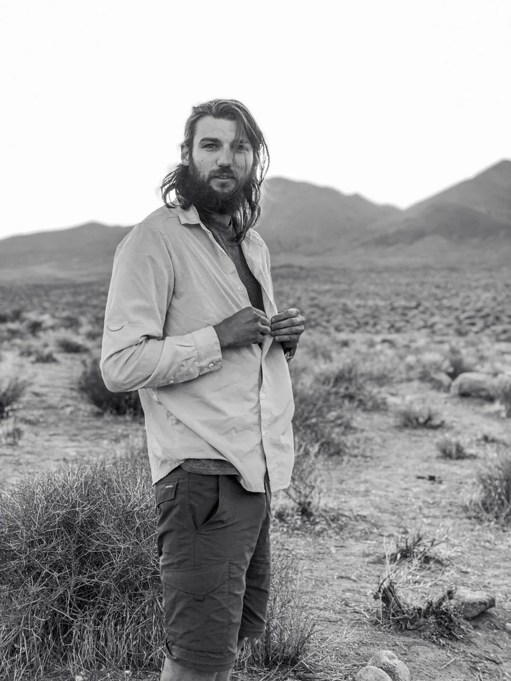 A man with long hair and a beard standing in a desert landscape with mountains in the background, looking at the camera.