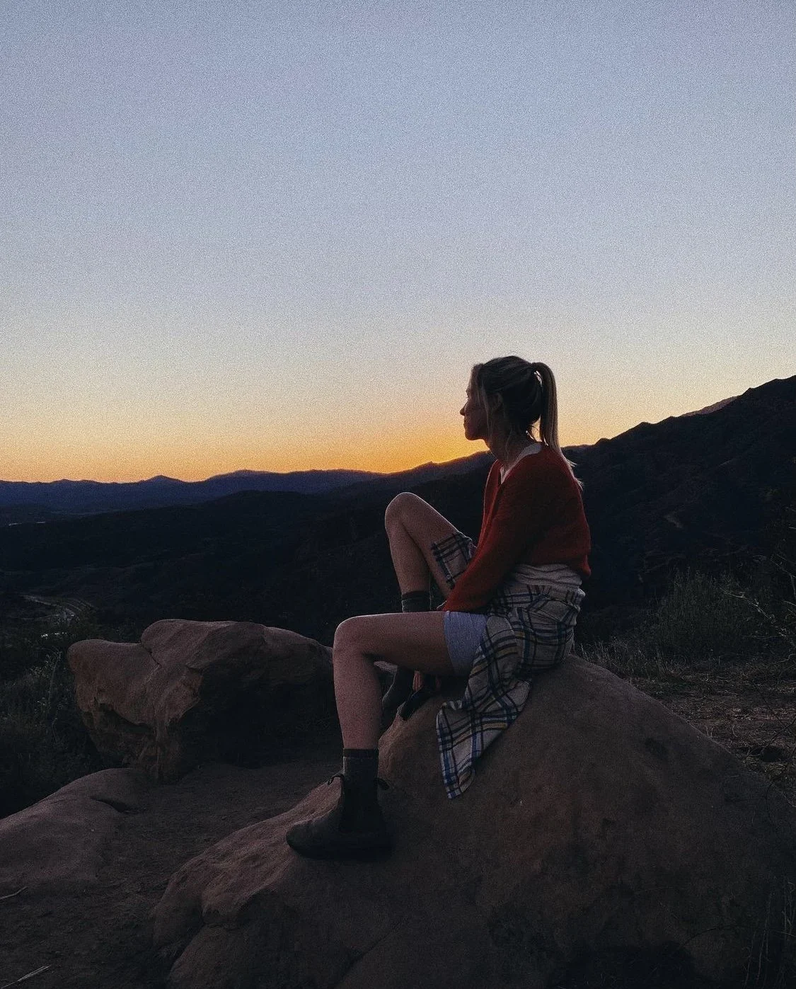 A person sitting on a rock during sunset, overlooking a mountainous landscape with a colorful sky.
