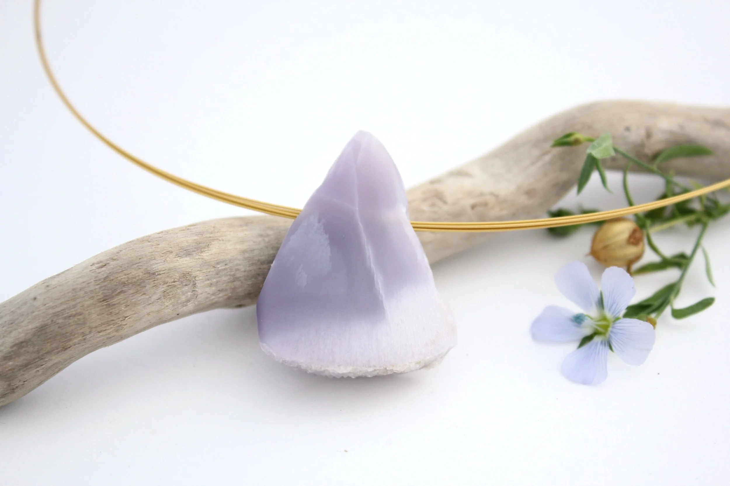 Un morceau de pierre violette, une branche de fleurs bleues et une branche de bois clair posés sur une surface blanche, avec un fil métallique doré en cercle autour.