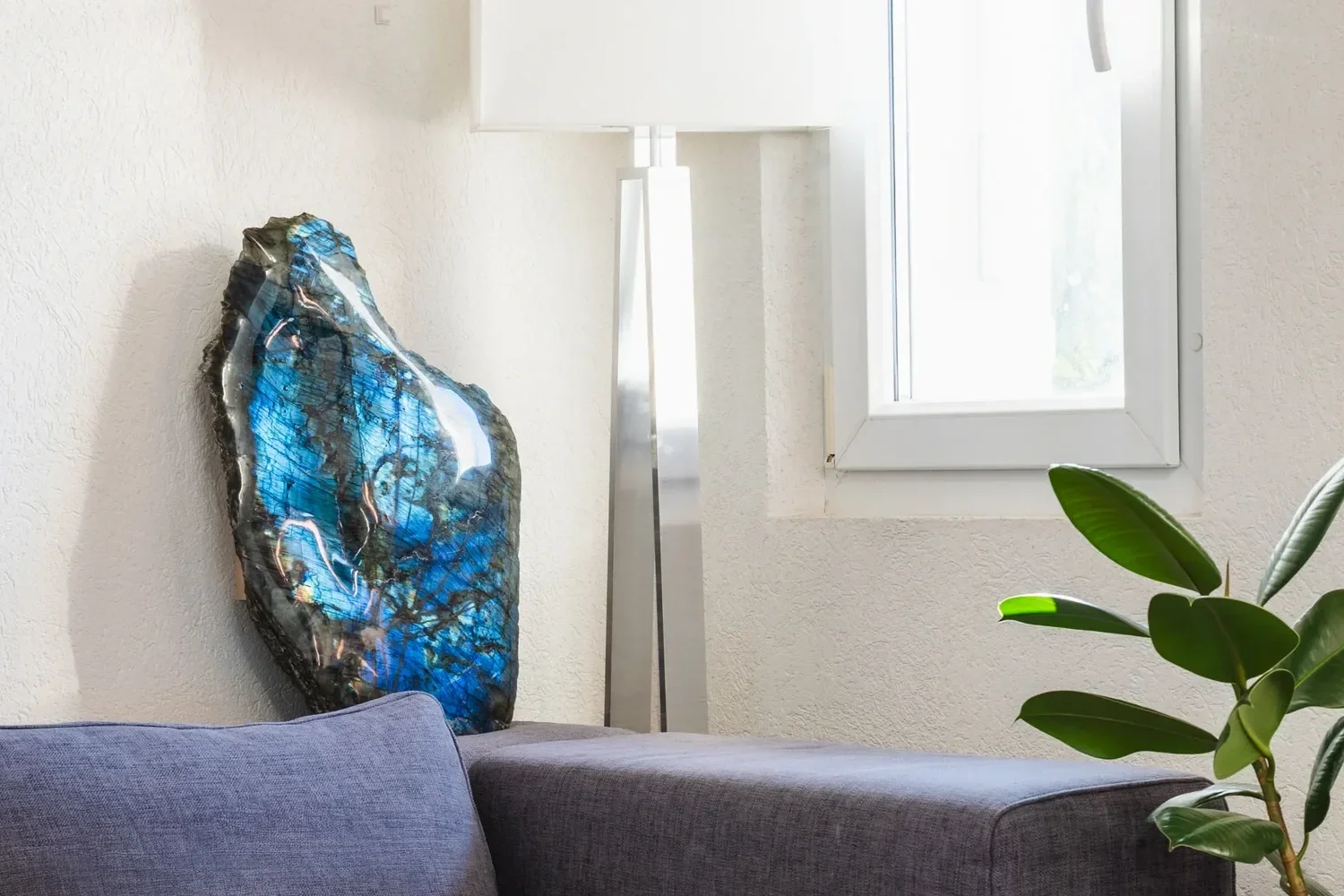 Intérieur d'un salon avec un coussin gris, une lampe de sol moderne en métal, un tableau en verre bleu et une plante verte près de la fenêtre