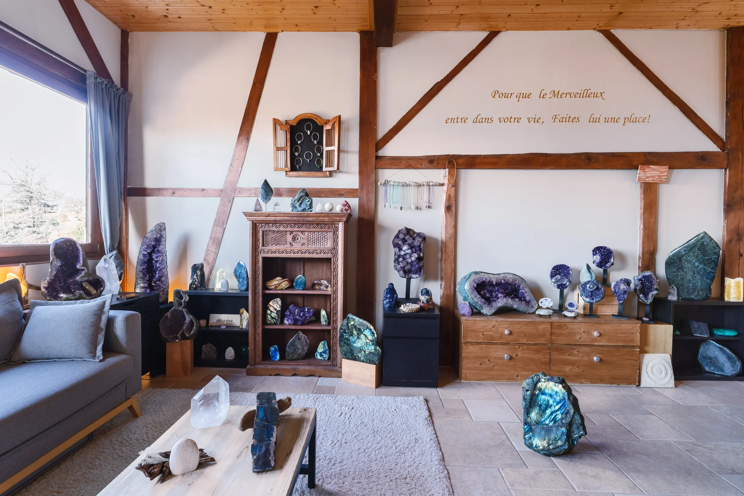 Une pièce décorée avec plusieurs cristaux et géodes en vitrine et sur des étagères en bois, avec un mur blanc comportant une citation en français.