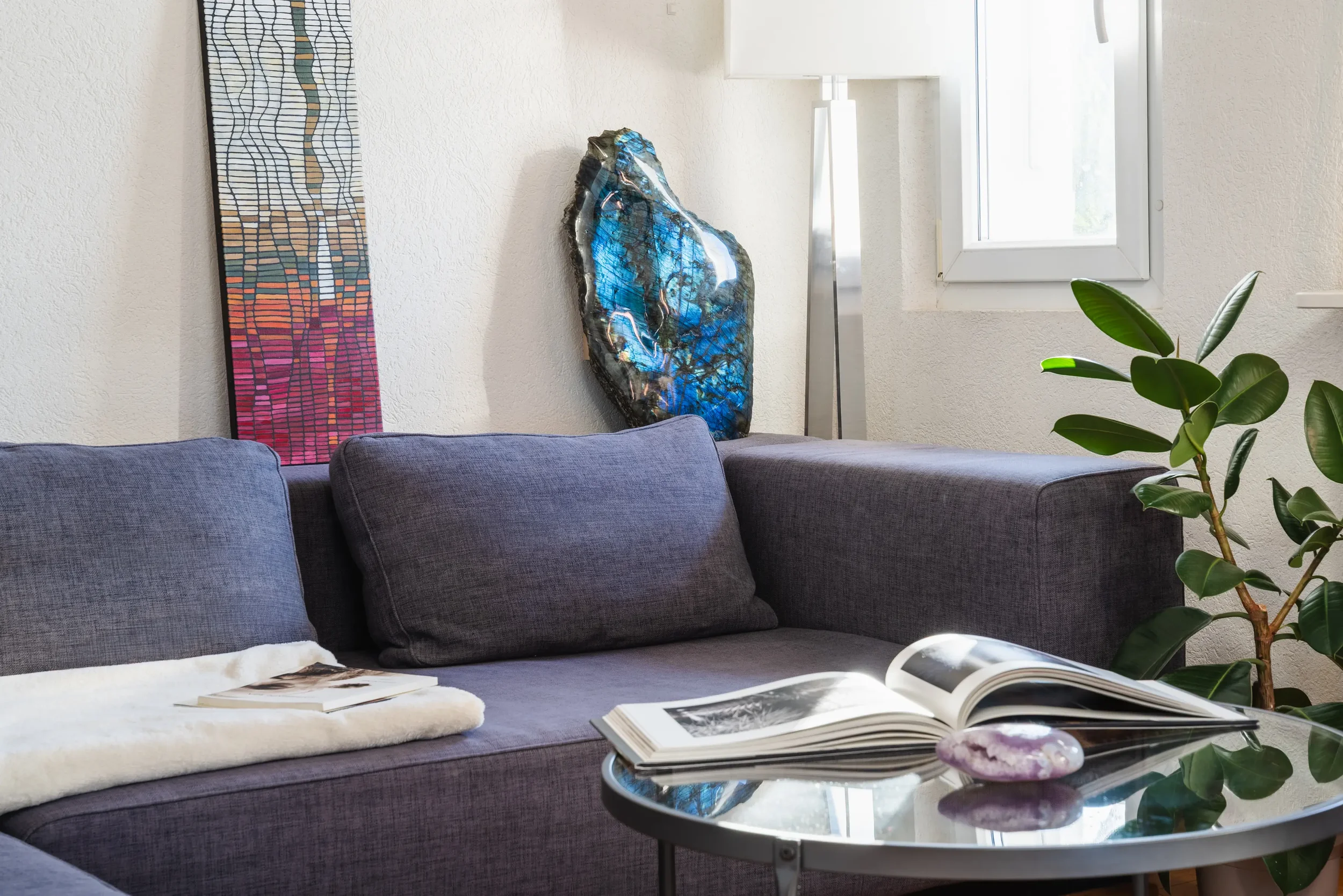 Salon avec un canapé gris, une table noir avec un livre ouvert et une pierre, une plante verte à droite, un tableau coloré et une roche décorative accroche sur le mur, une lampe et une fenêtre