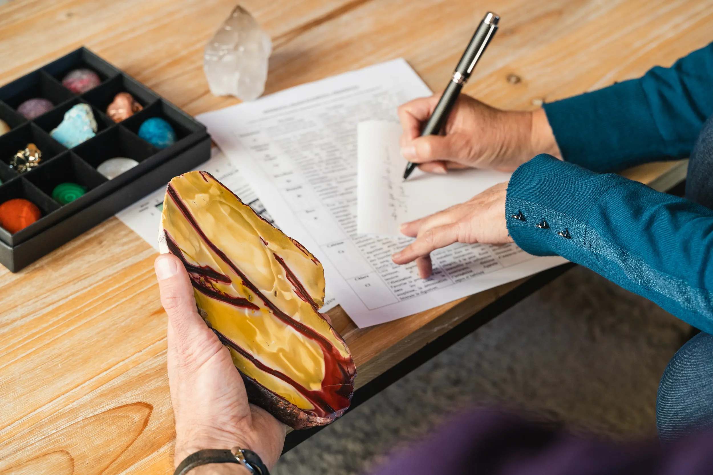 Une personne tient un gros caillou peint avec des motifs jaunes, rouges et noirs. Une autre personne écrit sur un formulaire posé sur une table en bois. Sur la table, il y a une boîte avec des pierres de différentes couleurs et un cristal clair.