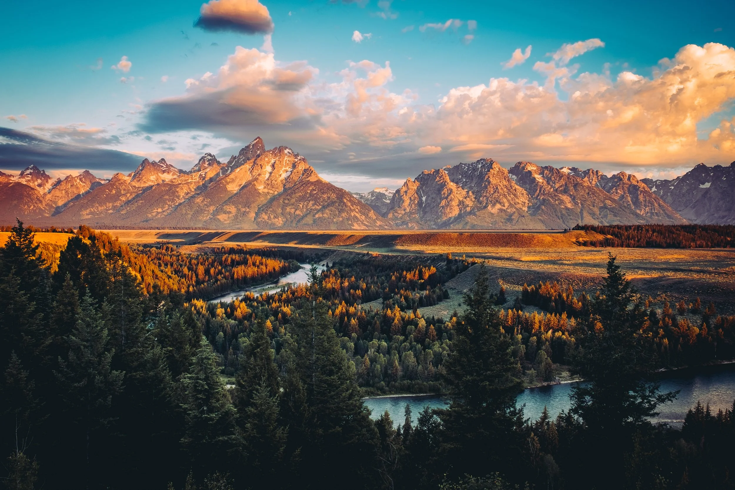Sunset over mountain range with pine forest, river, and colorful sky with clouds.