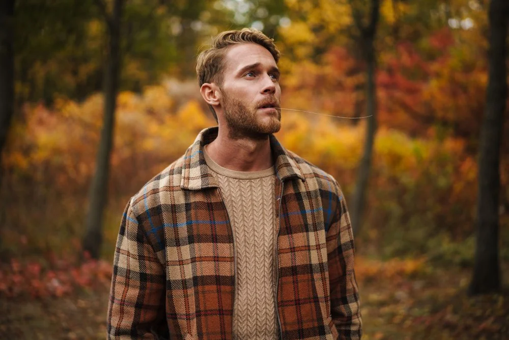 A man with a beard and light brown hair looking upwards while walking in a forest with fall foliage, wearing a plaid jacket and a beige cable-knit sweater.