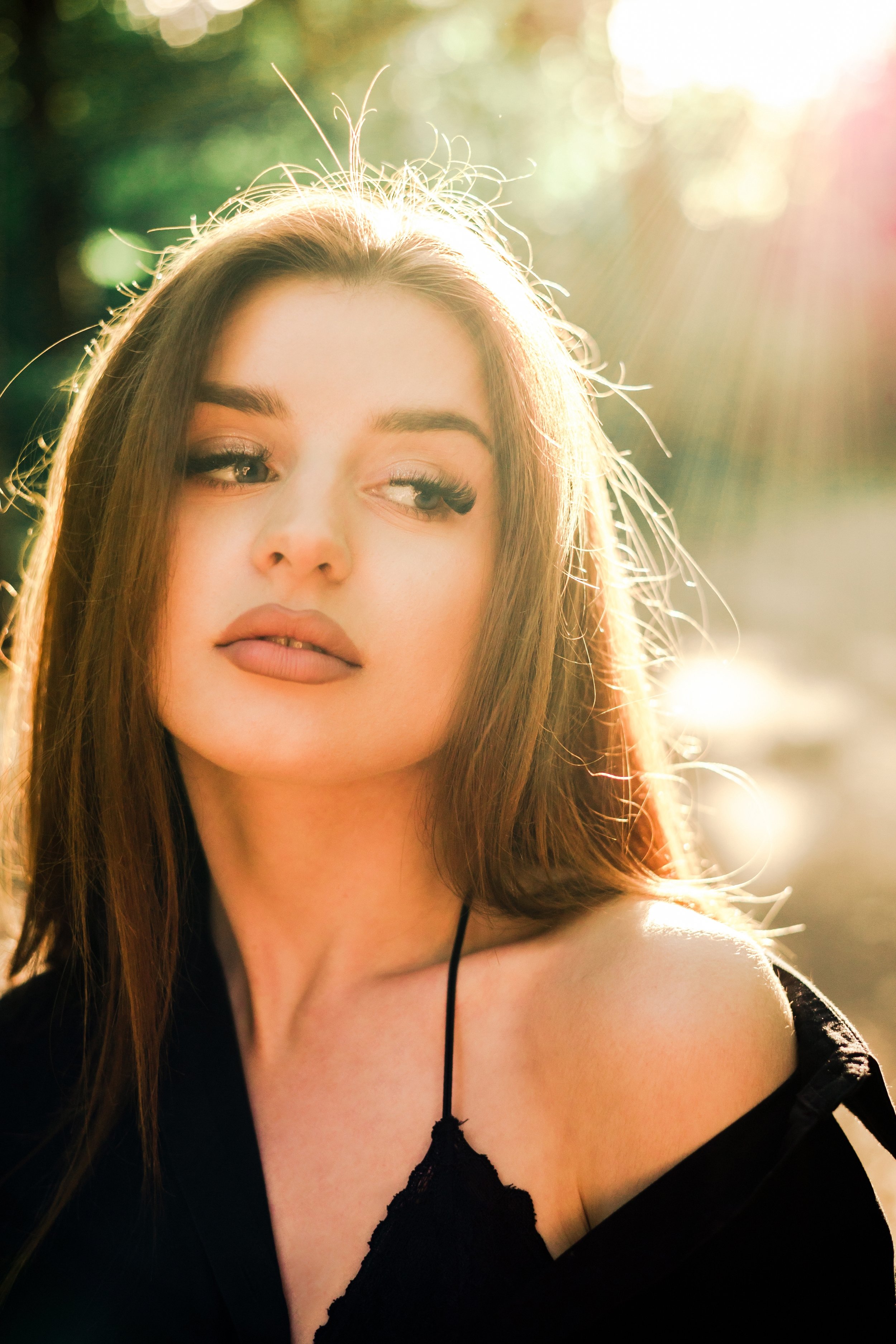 A young woman with long brown hair and makeup, looking to the side, with sunlight behind her creating a backlit effect.