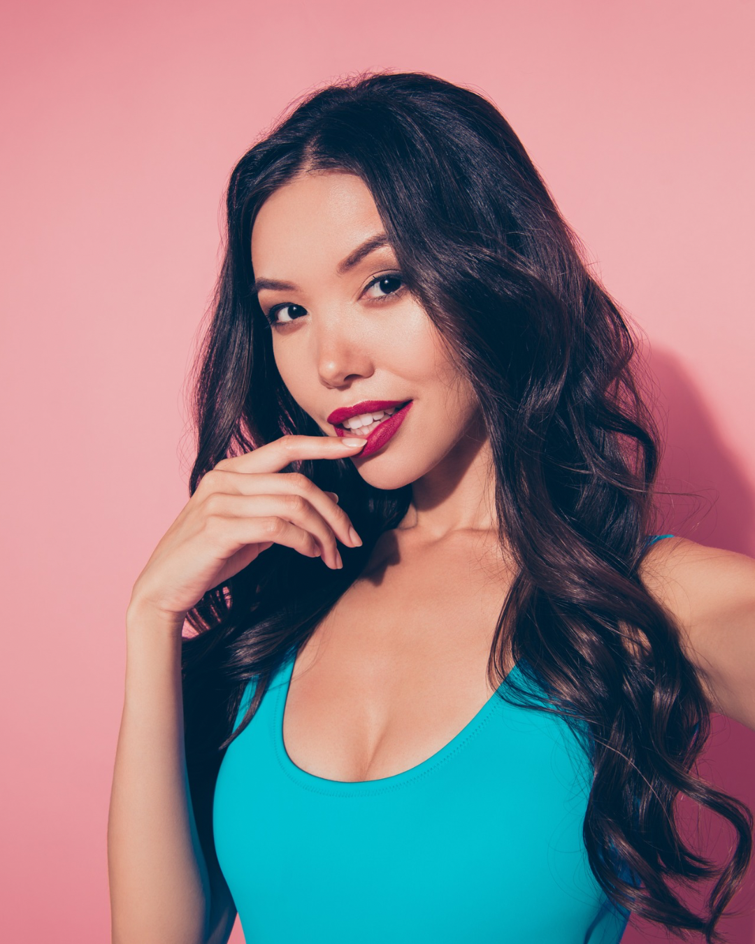 A young woman with long, dark, wavy hair wearing a bright blue top, red lipstick, and posing with her finger near her mouth against a pink background.