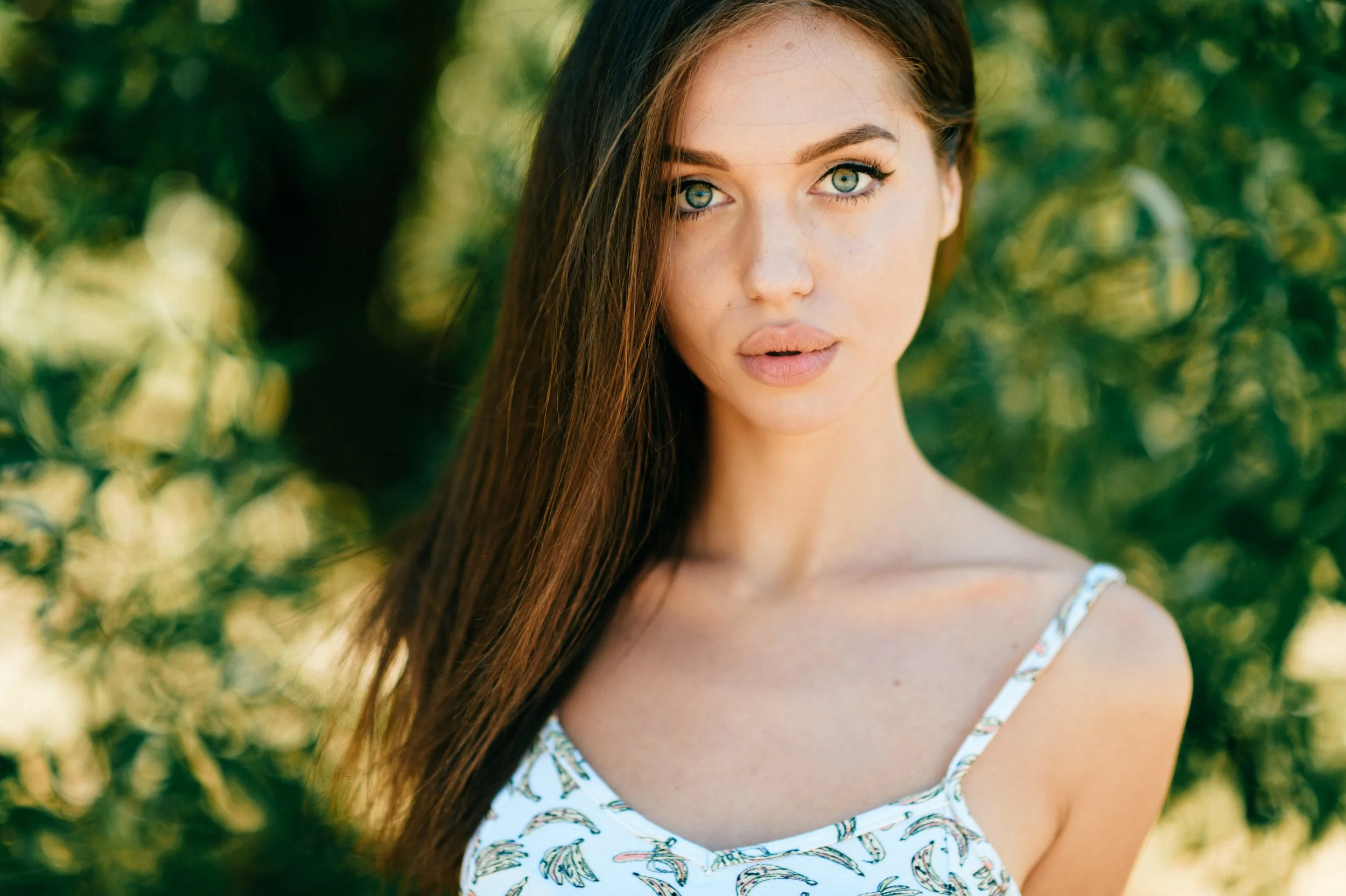 A young woman with long brown hair and striking blue eyes looking directly at the camera, wearing a white top with a palm tree pattern, outdoors with a background of green foliage.