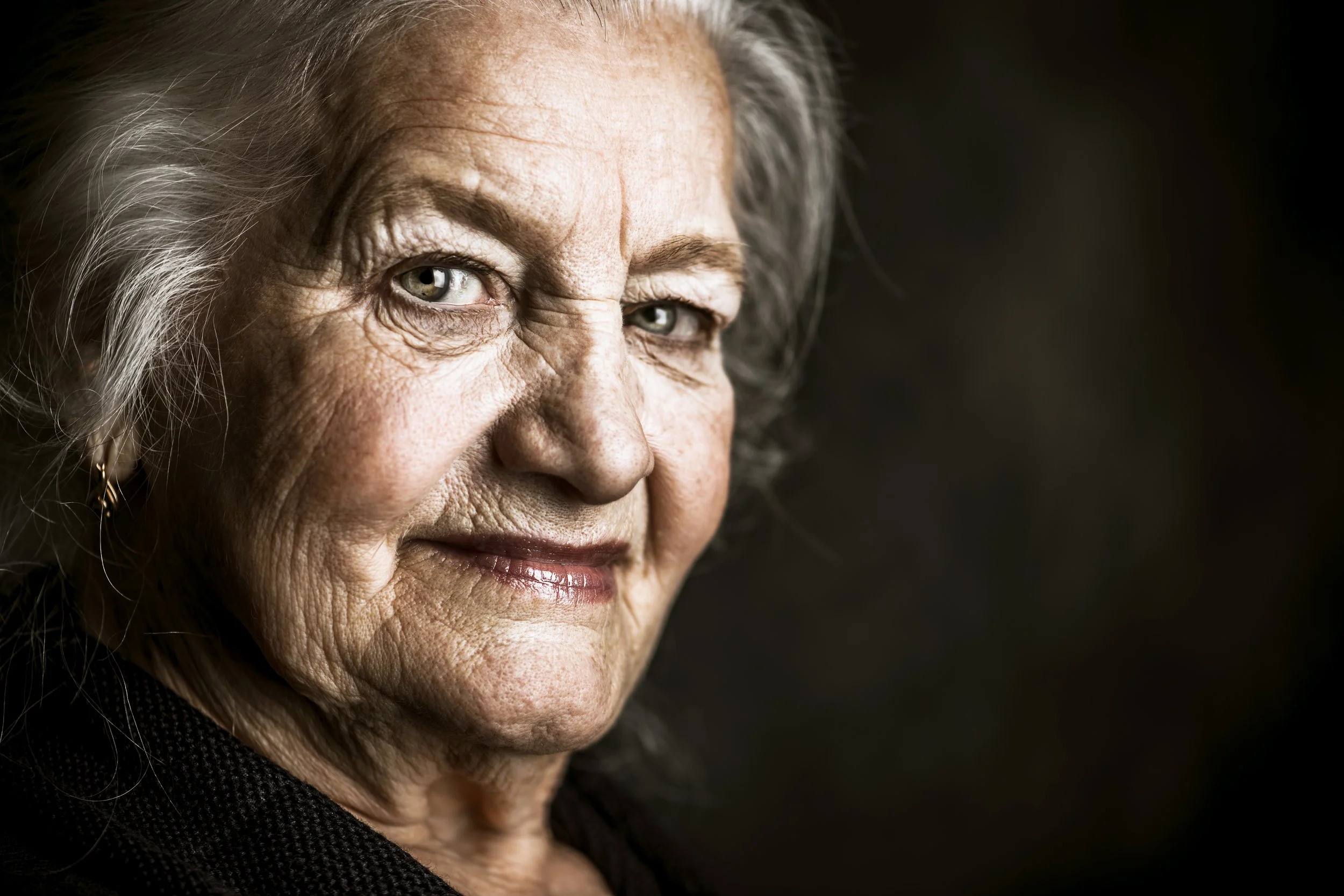 Close-up portrait of an elderly woman with gray hair, light-colored eyes, and a slight smile, wearing a black top and small earrings, against a dark background.